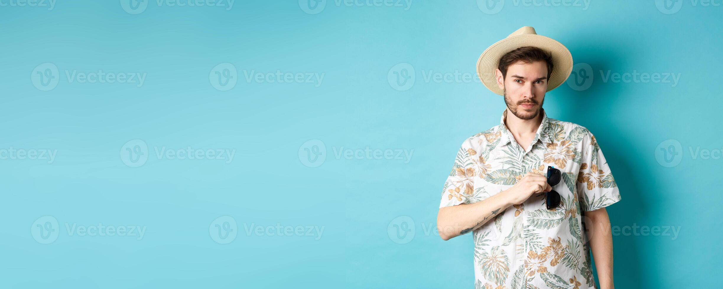 Handsome tourist in hawaiian shirt and summer hat, put sunglasses in pocket, going on vacation, standing against blue background photo
