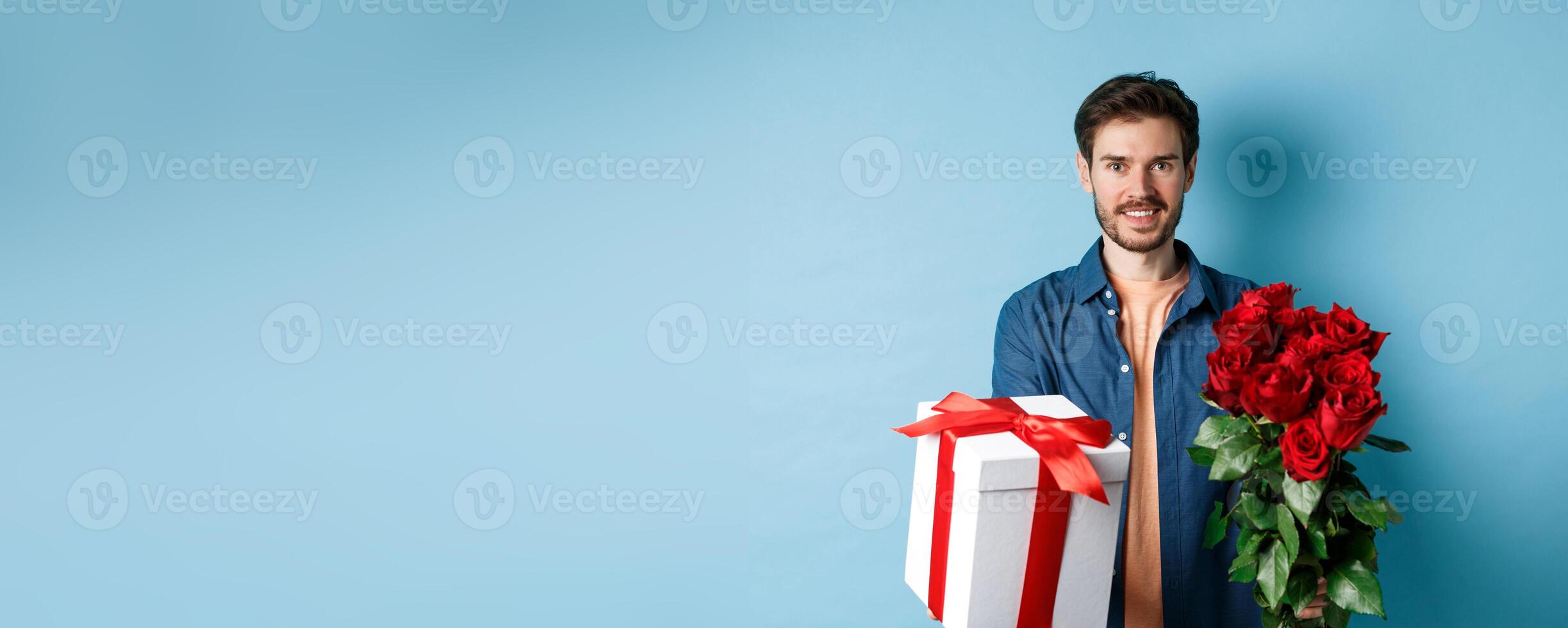 Love and Valentines day concept. Charming young man giving gift and bouquet of roses to girlfriend, standing over blue background photo