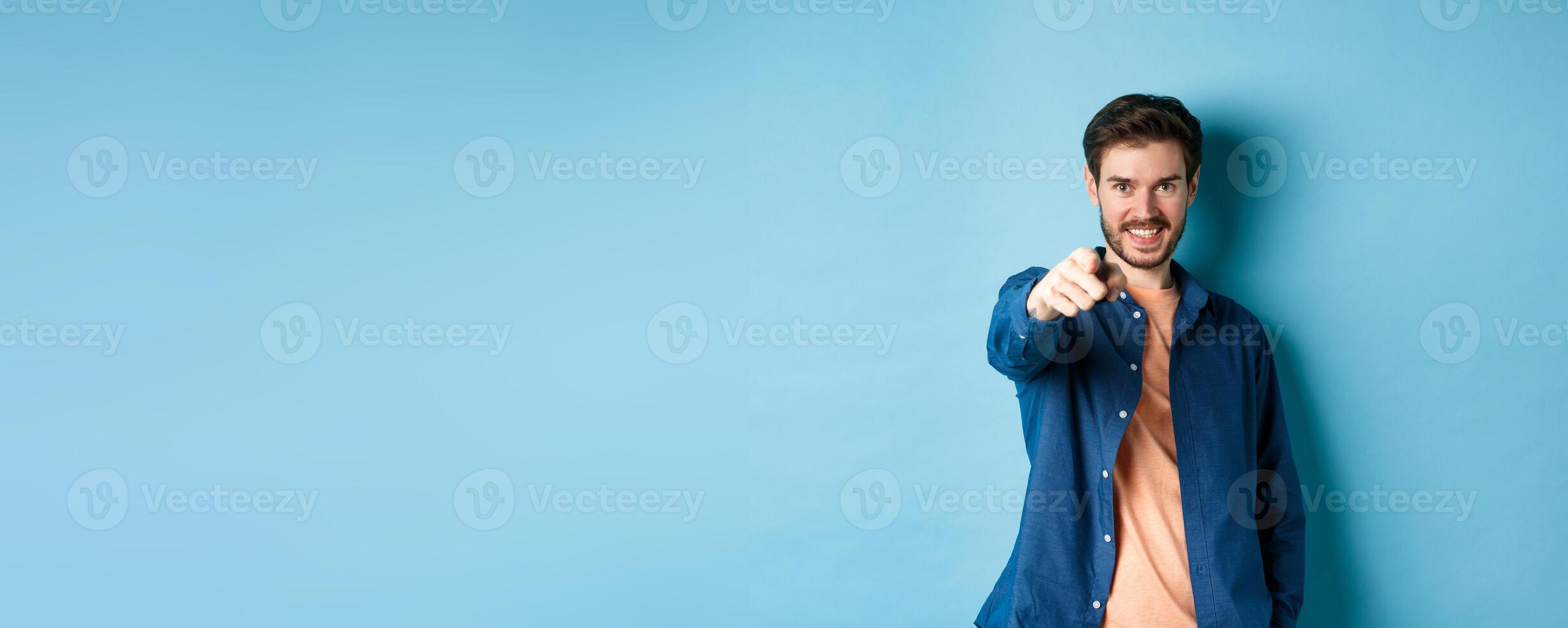 Confident smiling guy choosing or inviting you, pointing finger at camera decisive, standing on blue background photo