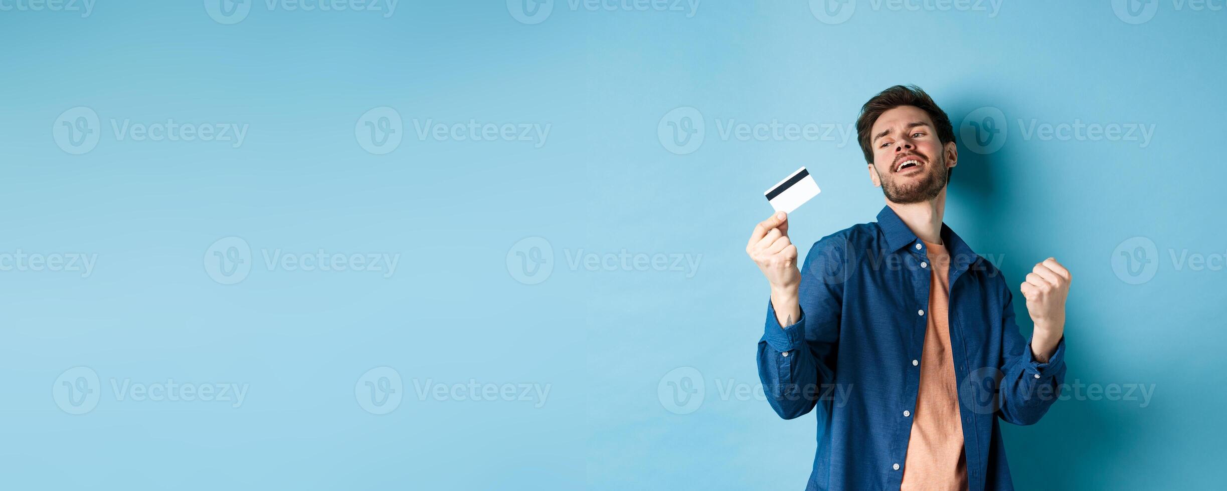Happy and satisfied young man dancing with plastic credit card, fist pump and smile, standing on blue background photo