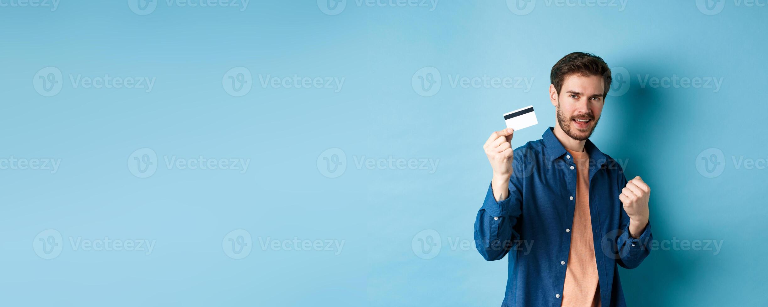 Satisfied young man showing plastic credit card and say yes, cheering of joy, standing on blue background photo