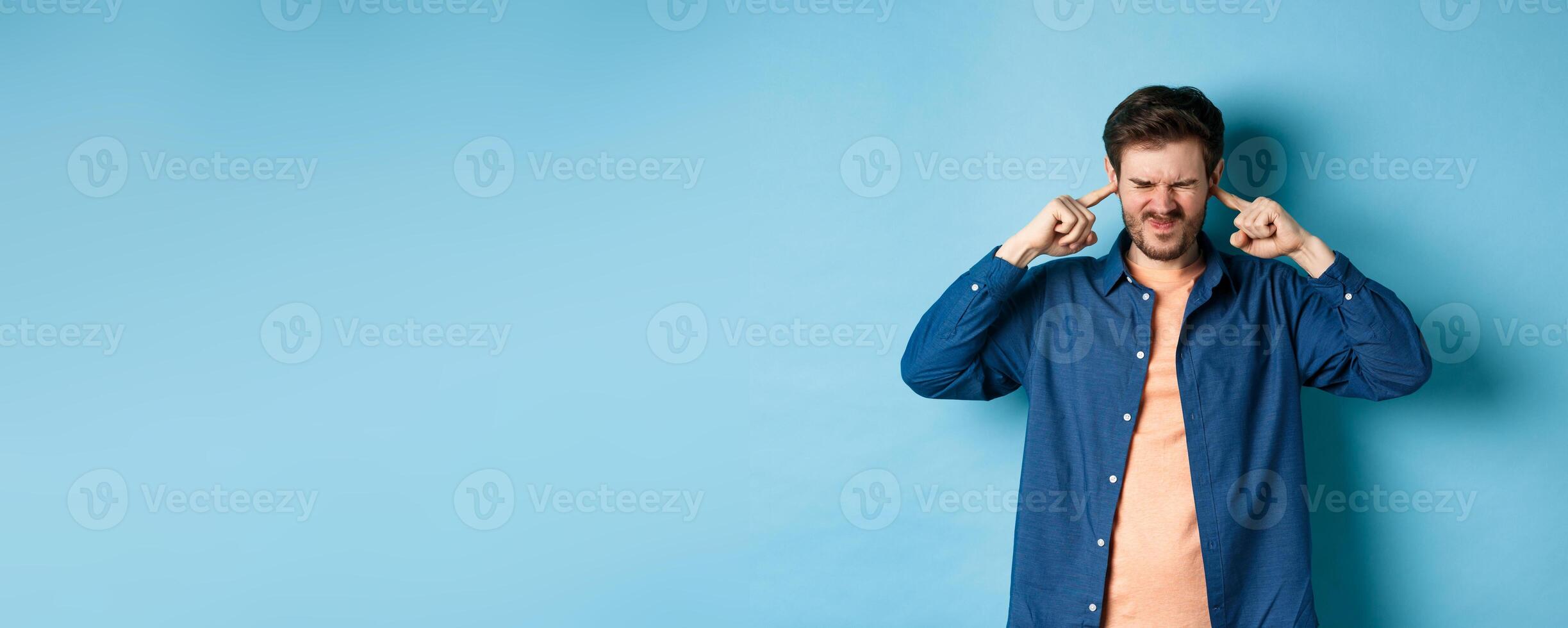 Annoyed young man block sound, shut ears with fingers and grimacing, disturbed by loud music, standing on blue background photo