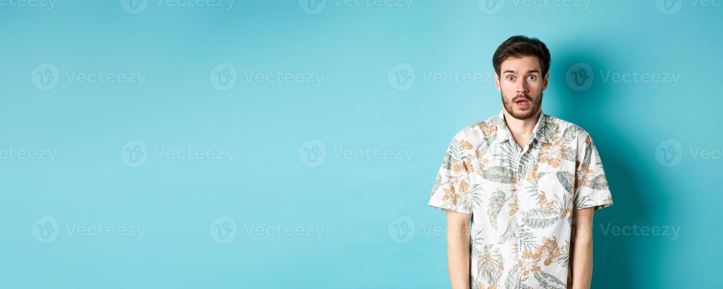 Image of shocked tourist guy drop jaw, gasping and looking startled, standing in hawaiian shirt on blue background photo