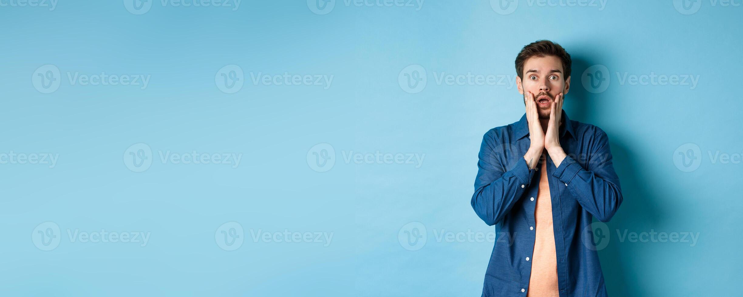Portrait of guy gasping and dropping jaw with shocked face, staring at camera startled, standing on blue background photo