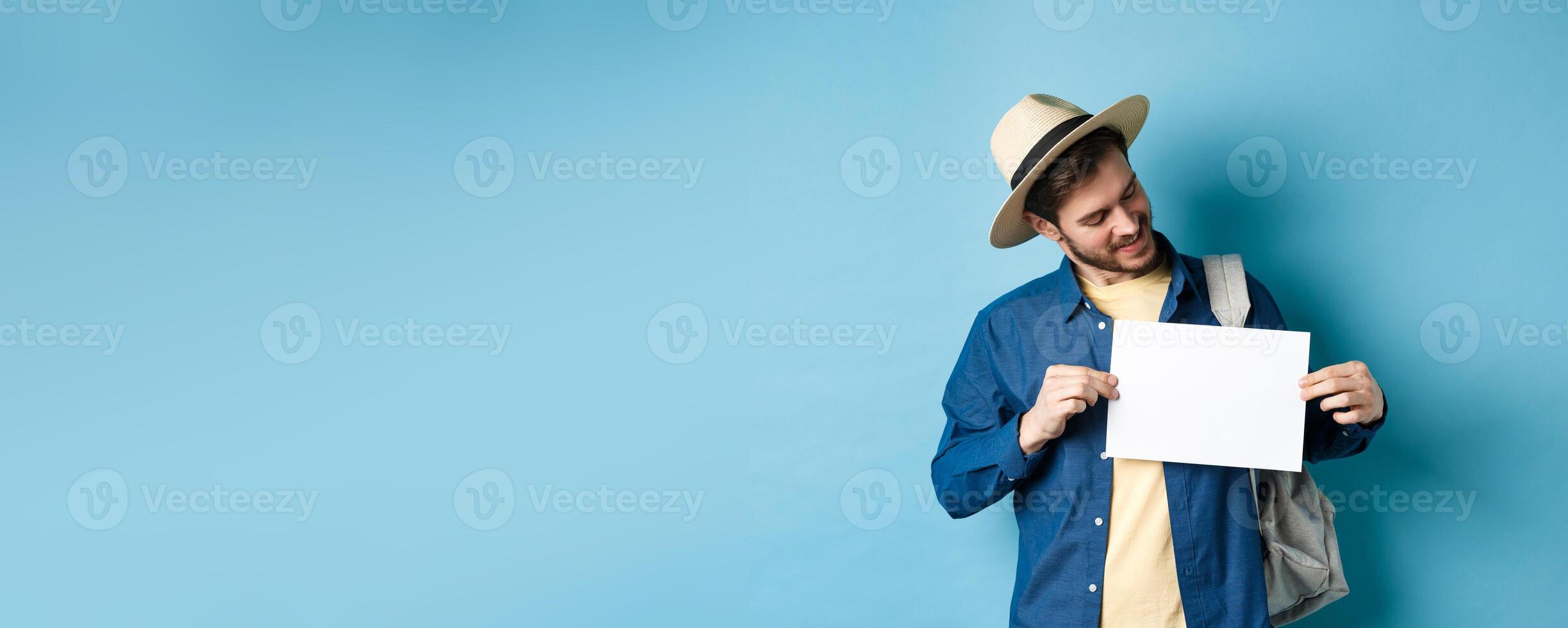 Happy tourist in straw hat smiling and looking at piece of paper for your logo, standing on blue background photo