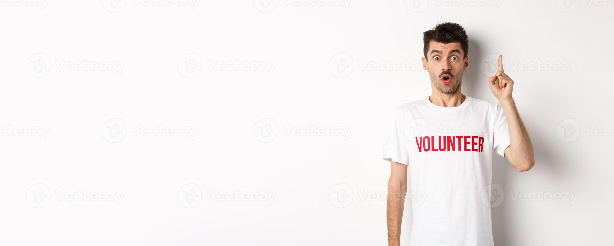 Handsome young man in volunteer t-shirt having an idea, raising finger and saying suggestion, pointing up, standing over white background photo