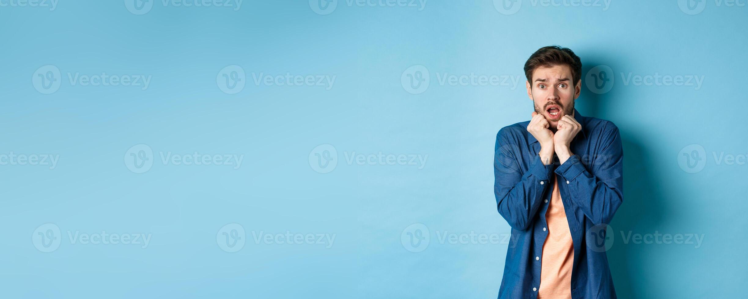 Scared young man gasping and screaming frightened, looking terrified at something, standing on blue background photo