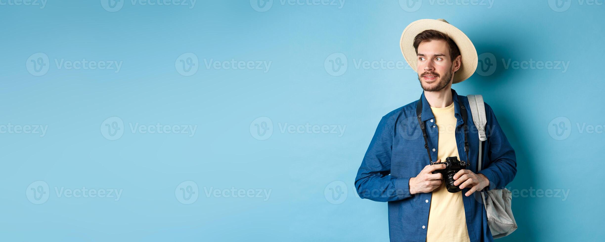 Handsome positive guy in summer hat, holding photo camera and looking aside, tourist taking pictures on vacation, standing with backpack on blue background