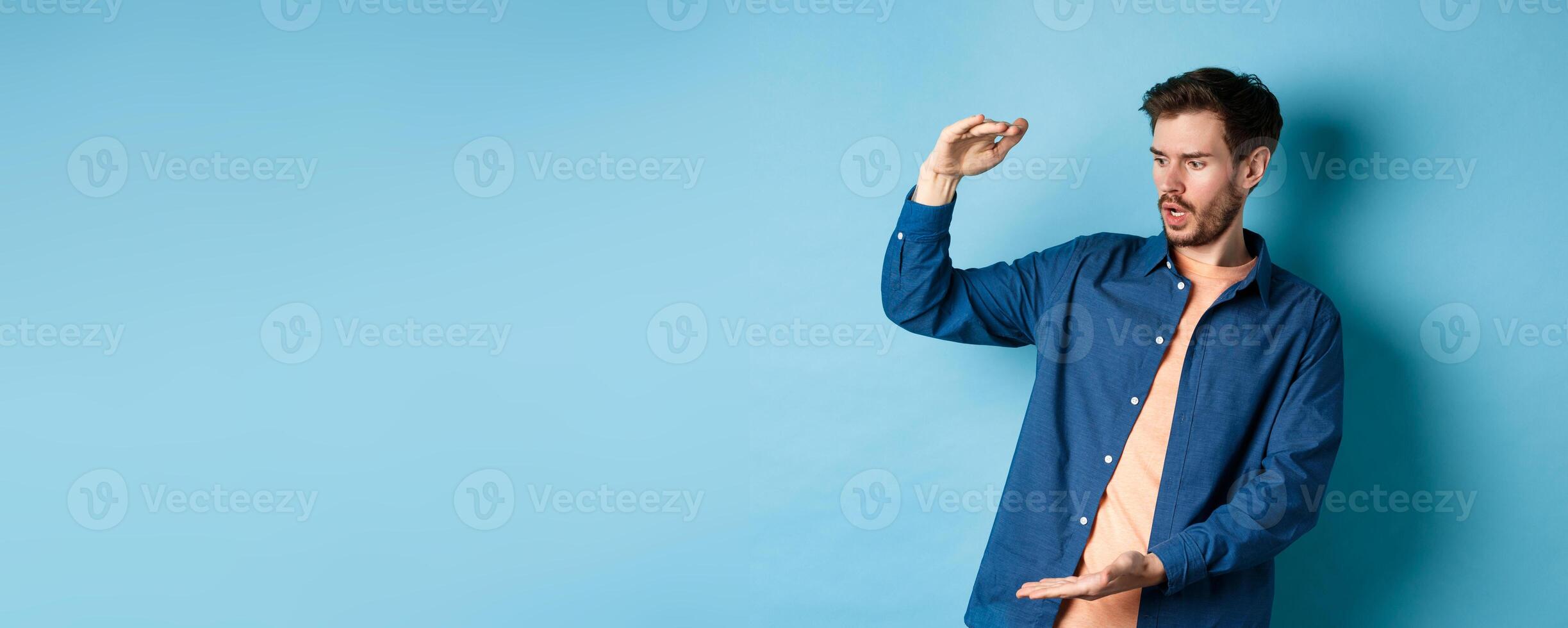 Shocked young man looking at empty space and showing big size object, standing on blue background photo