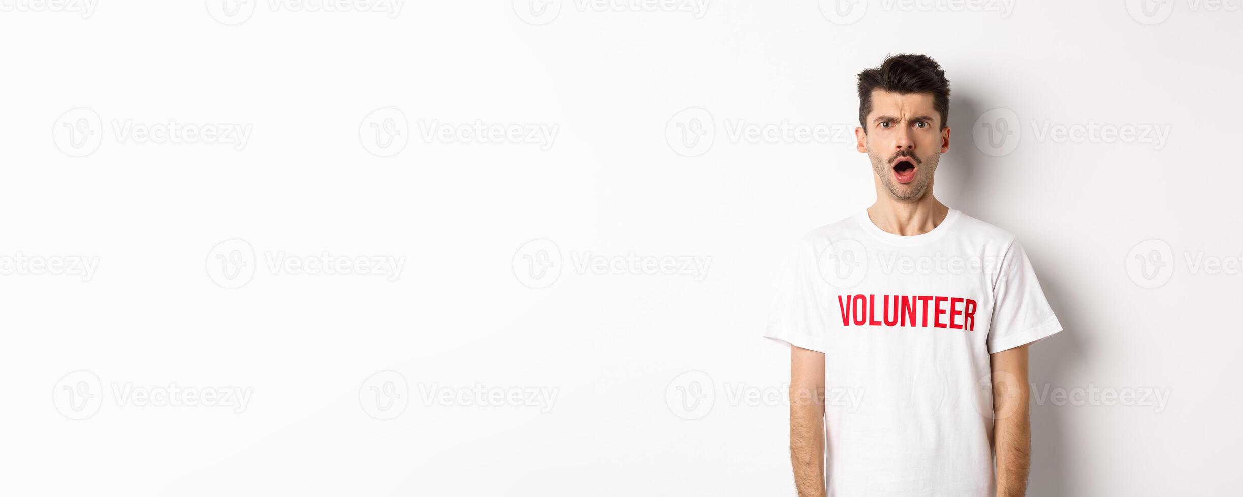 Shocked and confused man in volunteer t-shirt staring at camera and frowning displeased, standing against white background photo