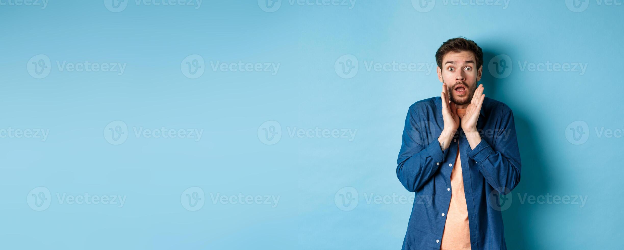 Shocked young man looking startled, screaming anxiously, standing on blue background photo