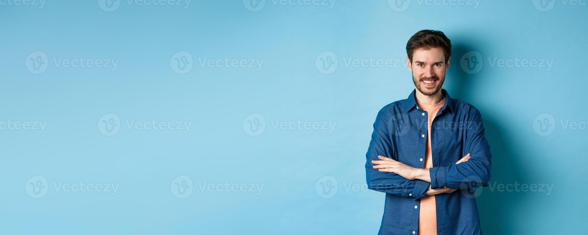 Confident young man feeling like professional, cross arms on chest and smiling sassy, standing on blue background photo