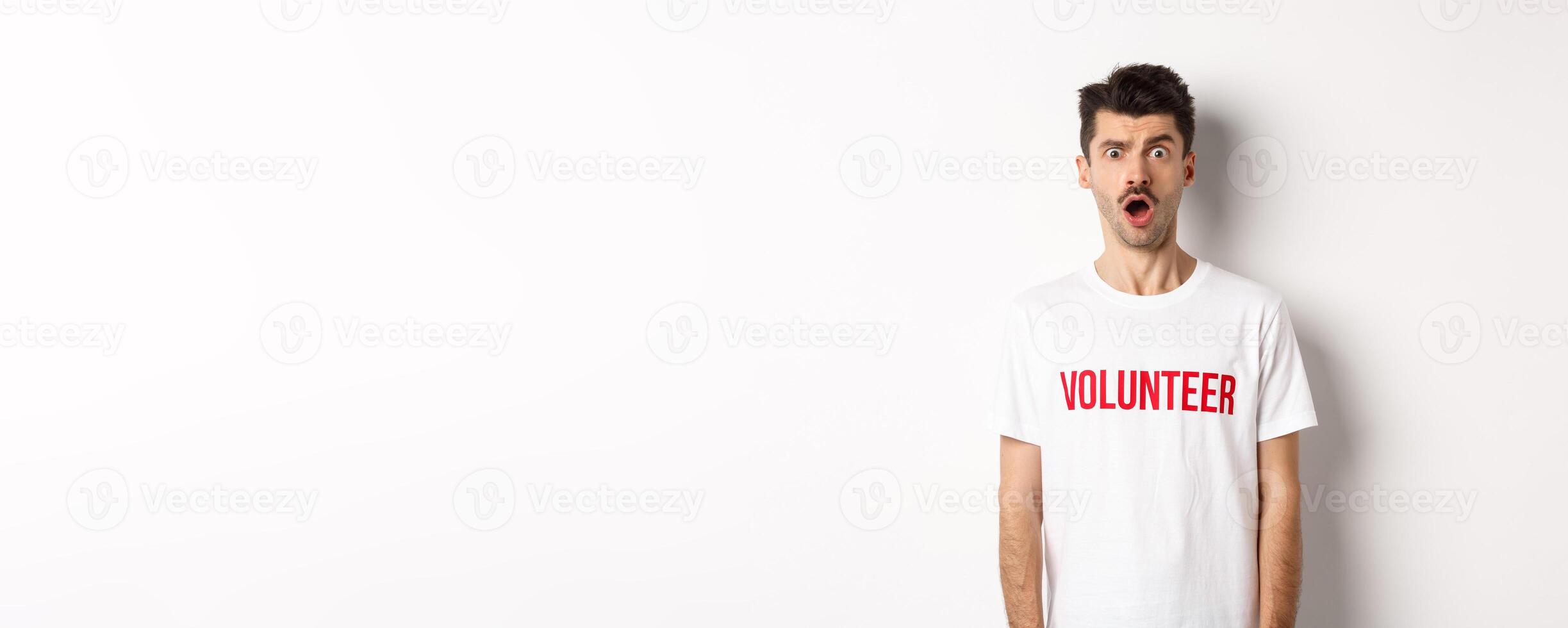 Shocked and confused man in volunteer t-shirt staring at camera speechless, standing against white background photo