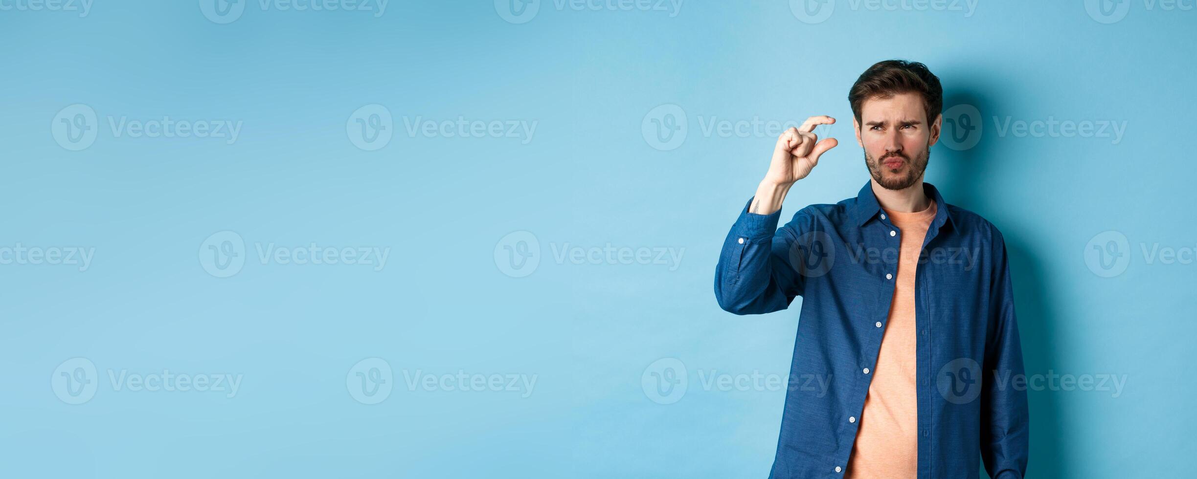Confused guy showing small size and looking doubtful or skeptical, standing over blue background photo
