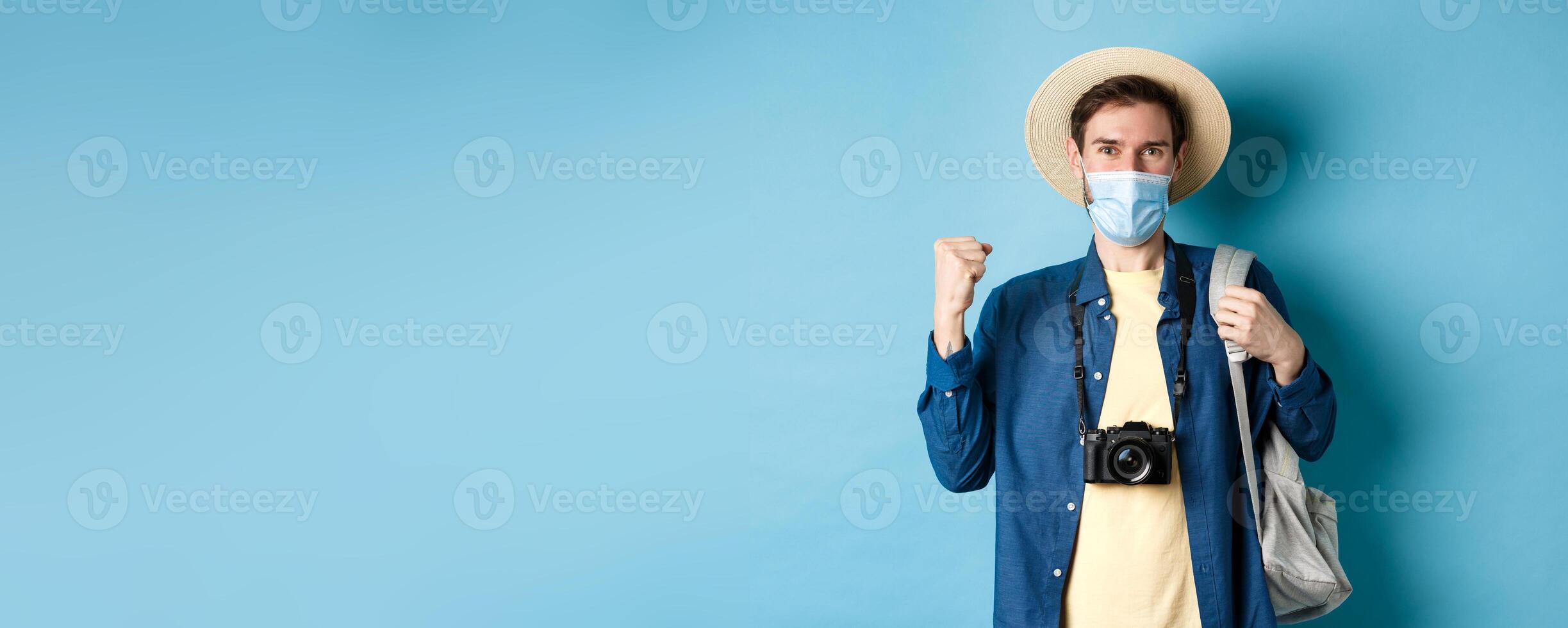 Covid-19 and summer holidays concept. Happy young man travelling abroad in medical mask, saying yes and raising fist up joyfully, cheerful about vacation, blue background photo