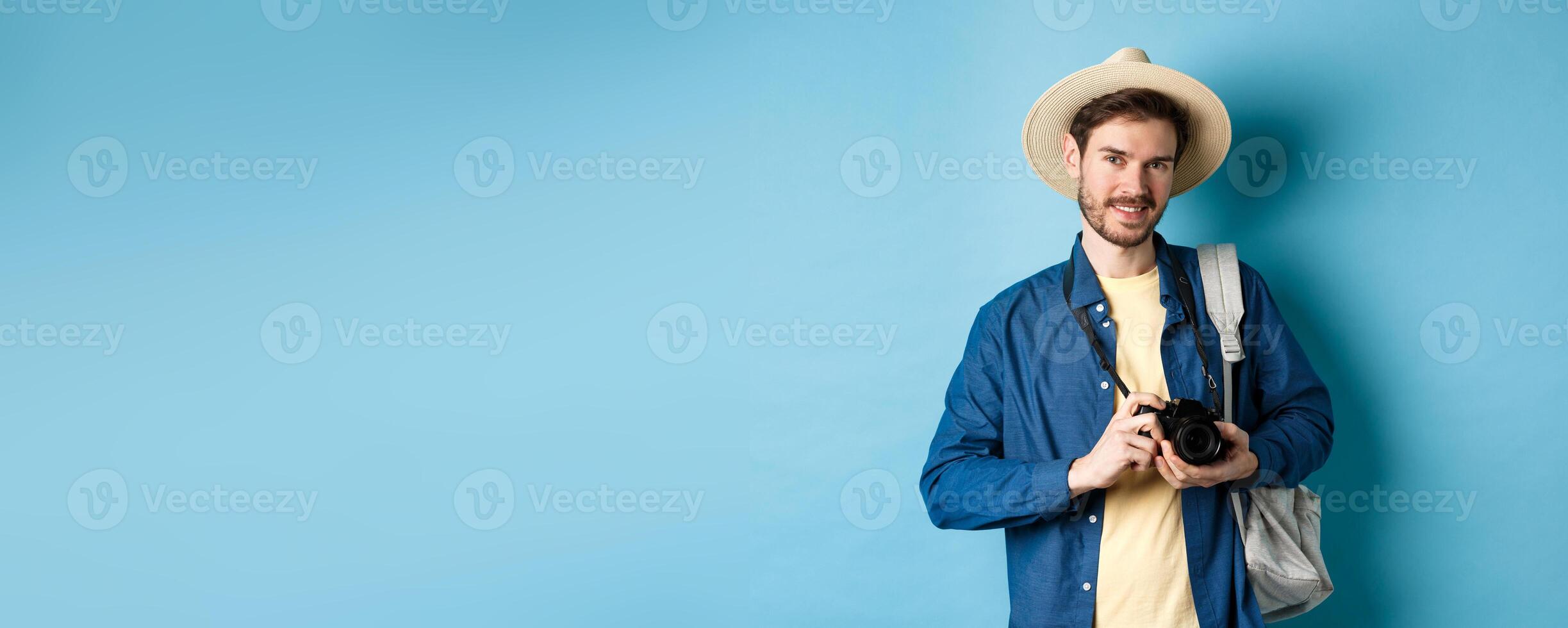 Handsome guy going on summer vacation, backpacking on holiday. Tourist with straw hat and camera smiling happy, standing on blue background photo