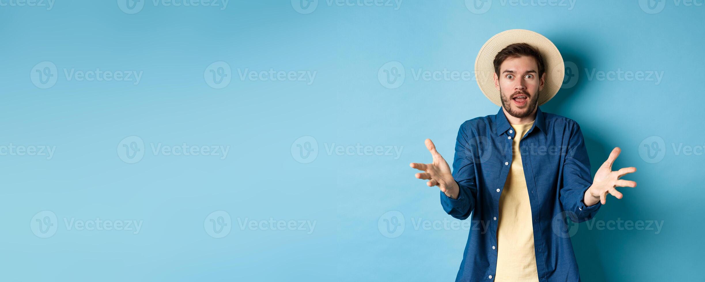 Surprised young man in summer hat, raising hands up staring with disbelief and amazement at camera, impressed with big event, standing on blue background photo
