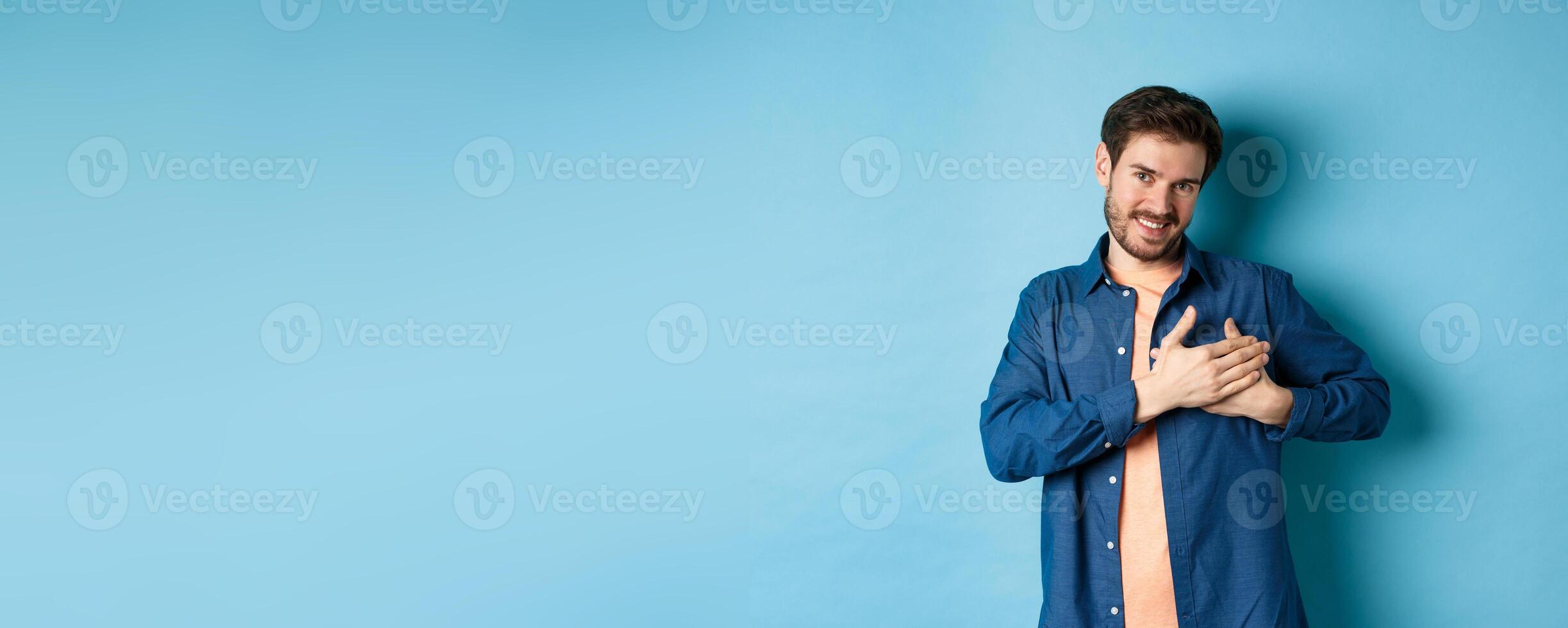 Smiling man feeling touched and grateful, holding hands on heart and thanking, standing on blue background photo
