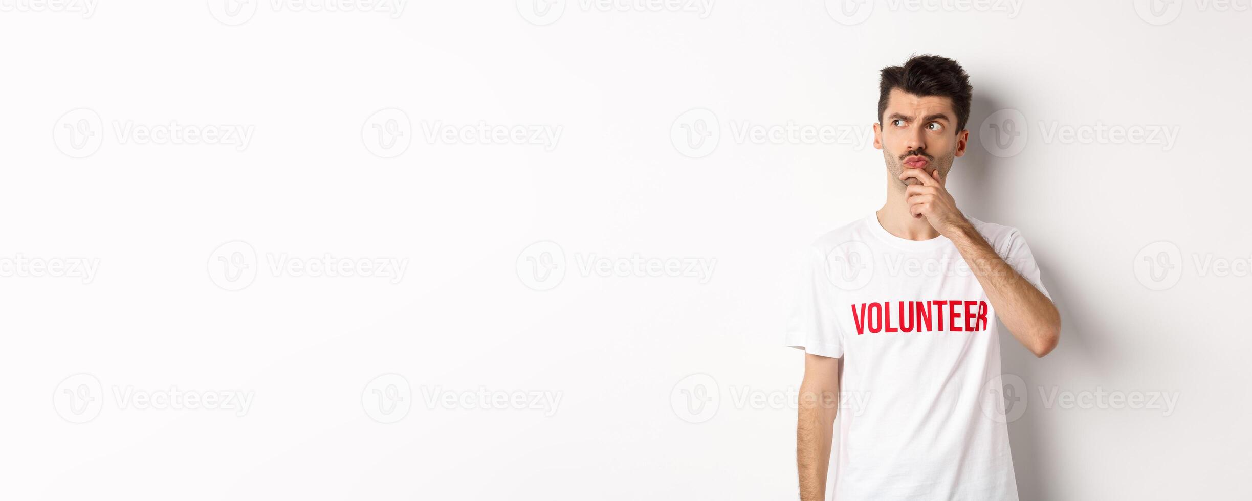 Thoughtful young male volunteer in white t-shirt searching solutions, looking at upper left corner and thinking, standing over studio background photo