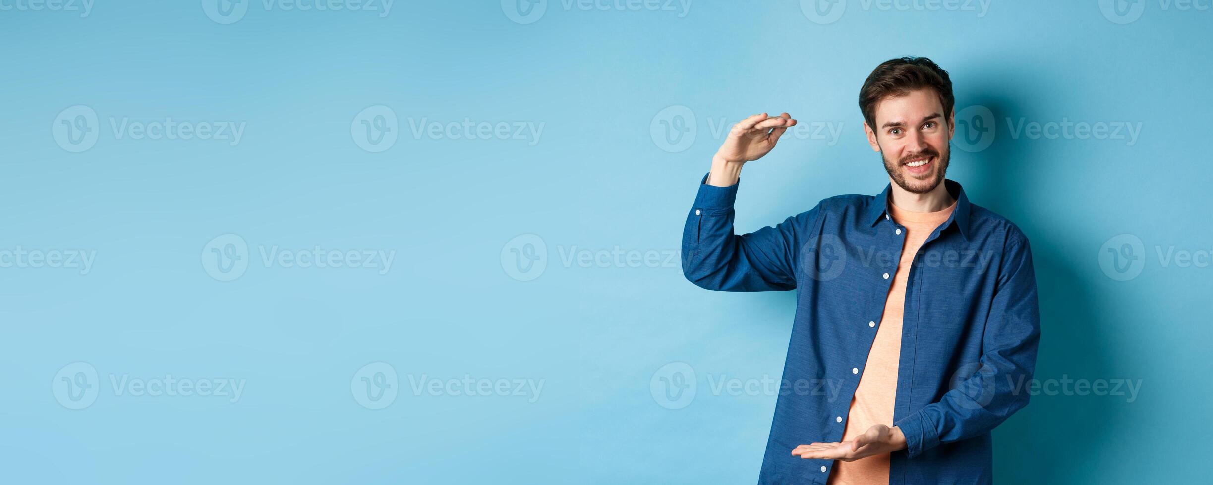 Handsome caucasian guy showing something big, shaping large size object with hands and smiling, standing on blue background photo