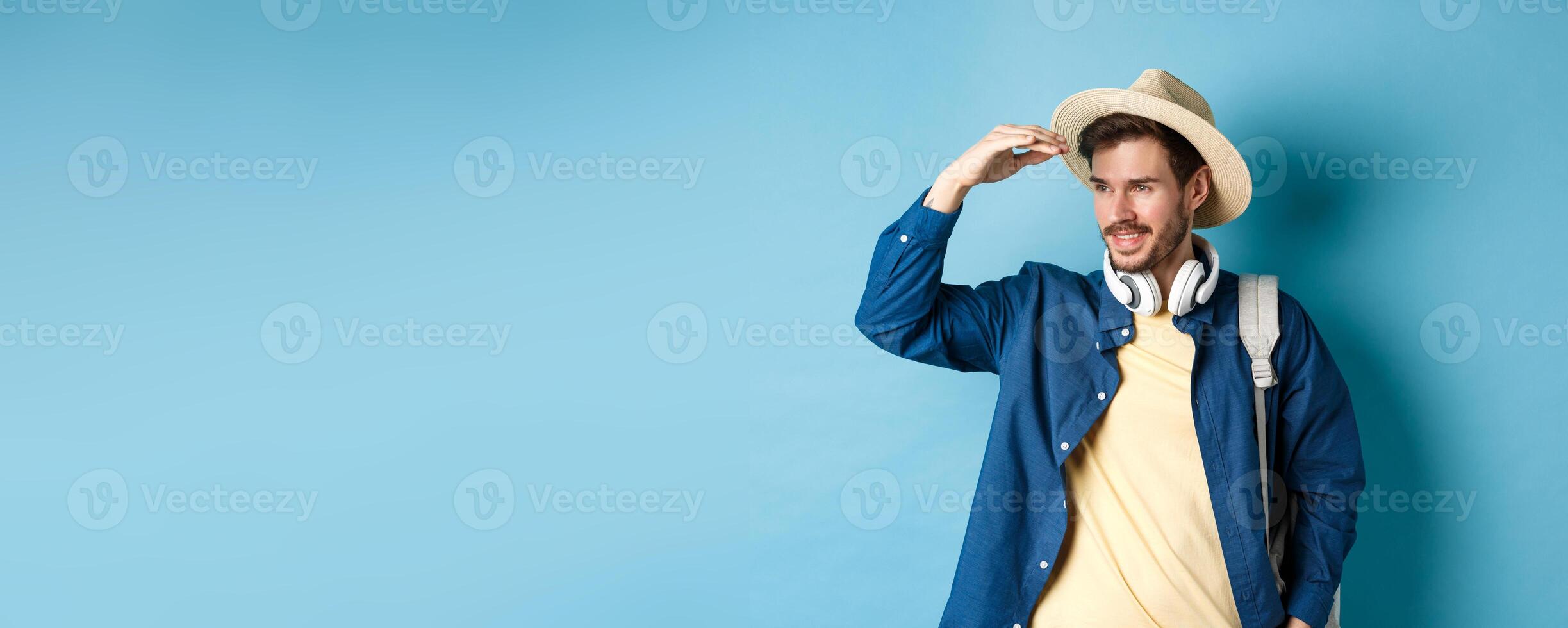 Handsome tourist in straw hat, covering face from sun and looking at distance aside, smiling pleased, travelling on summer vacation, standing over blue background photo
