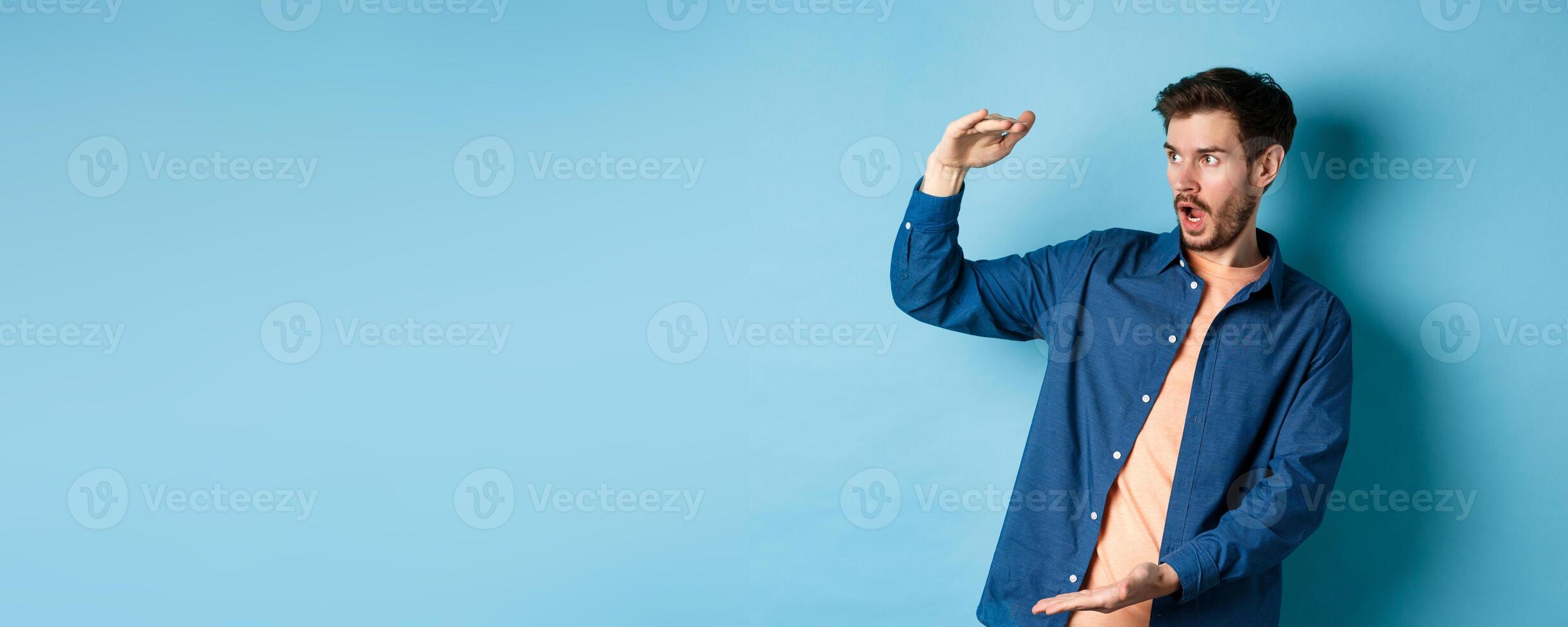 Impressed caucasian guy staring at empty space with big object, shaping large size, standing on blue background photo