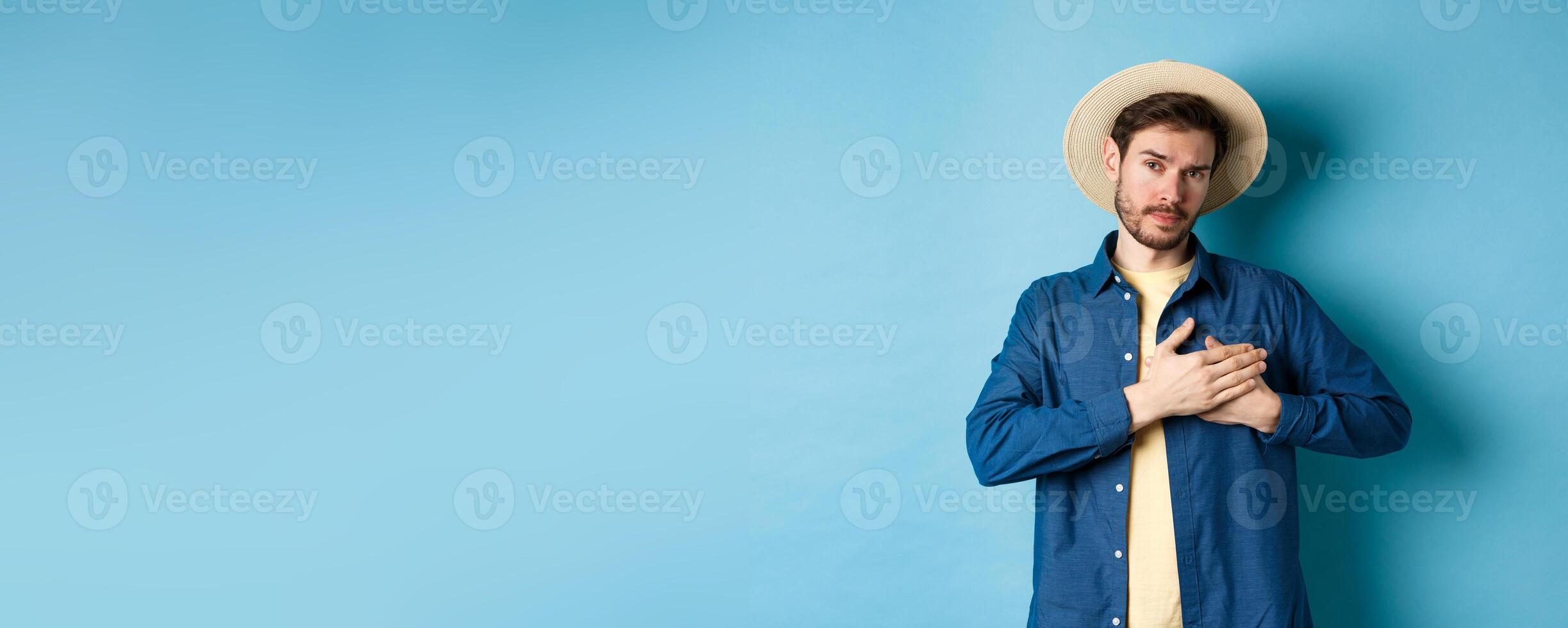 Heartfelt young woman in tourist hat, holding hands on heart and looking thankful and serious, standing on blue background photo