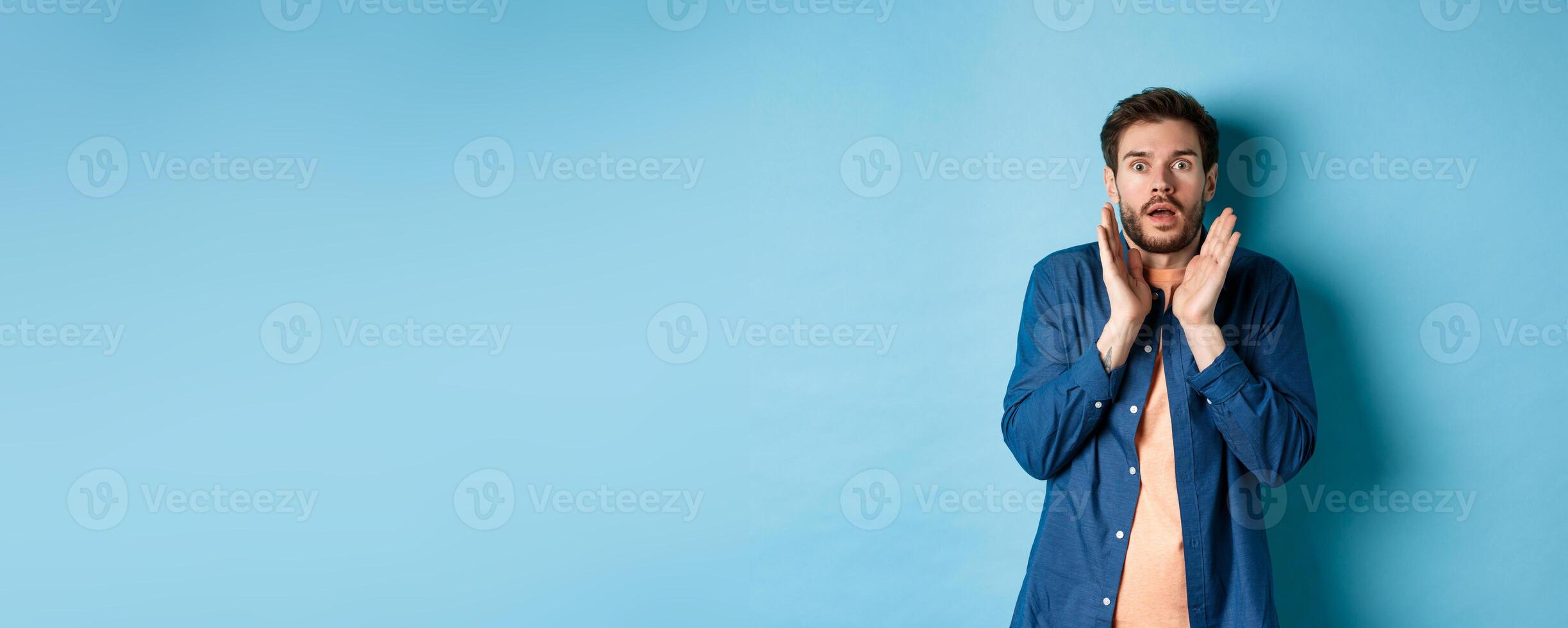 Shocked and scared young man standing in stupor, raising hands up and gasping speechless, standing on blue background photo
