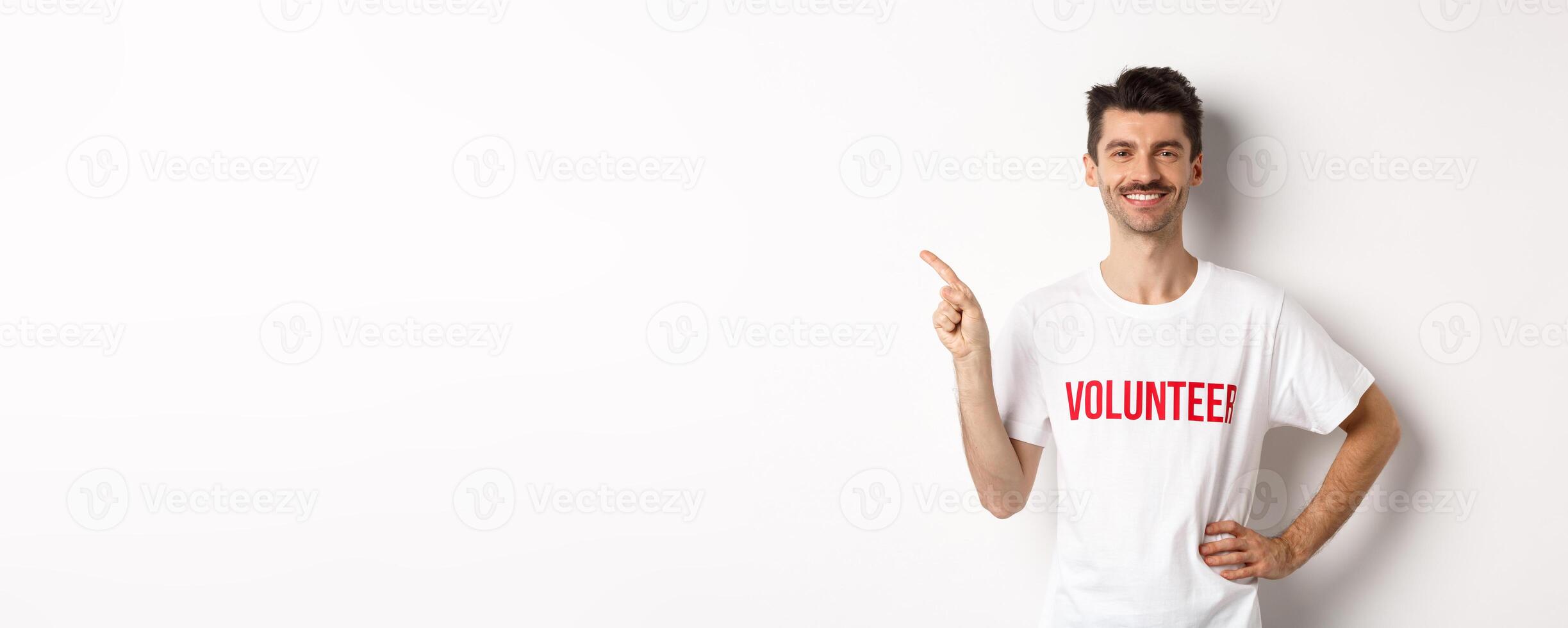 Handsome smiling volunteer in t-shirt pointing finger left, showing announcement, standing over white background photo