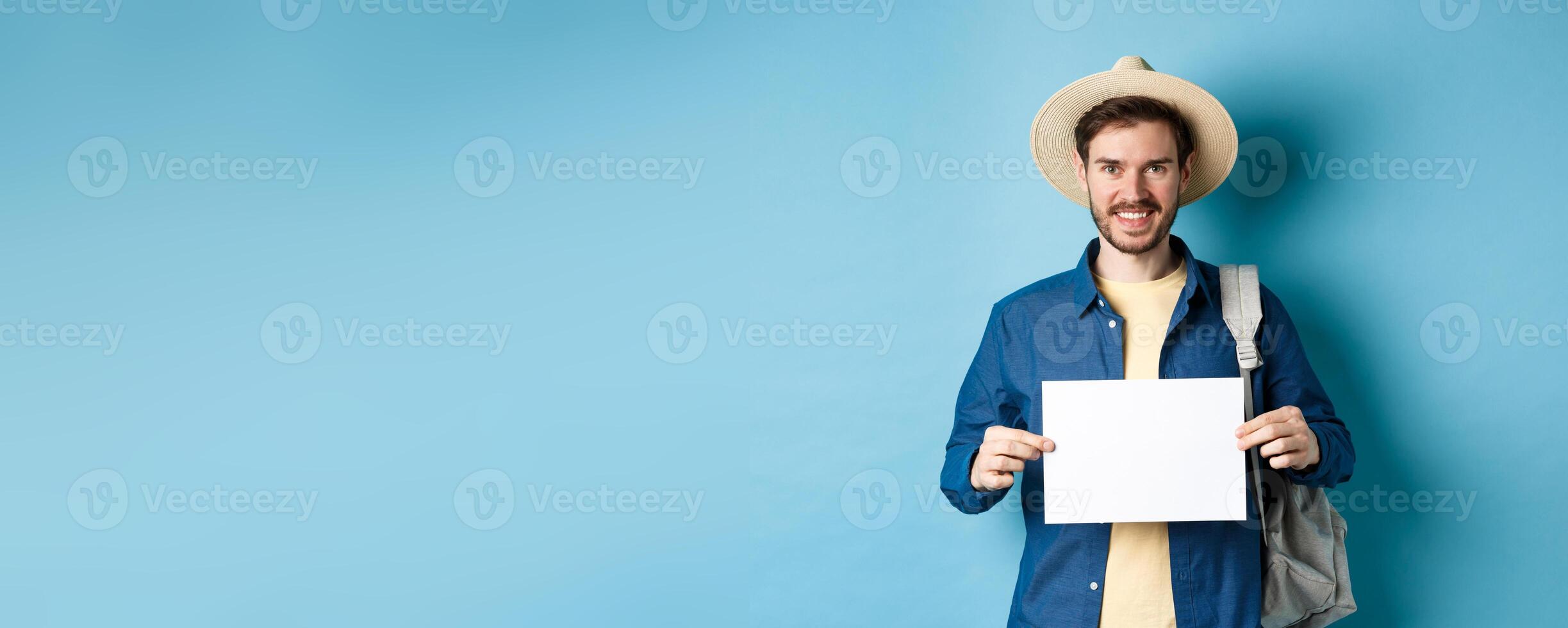Happy tourist with backpack showing empty piece of paper, smiling at camera, standing on blue background. Concept of summer holiday and vacation photo
