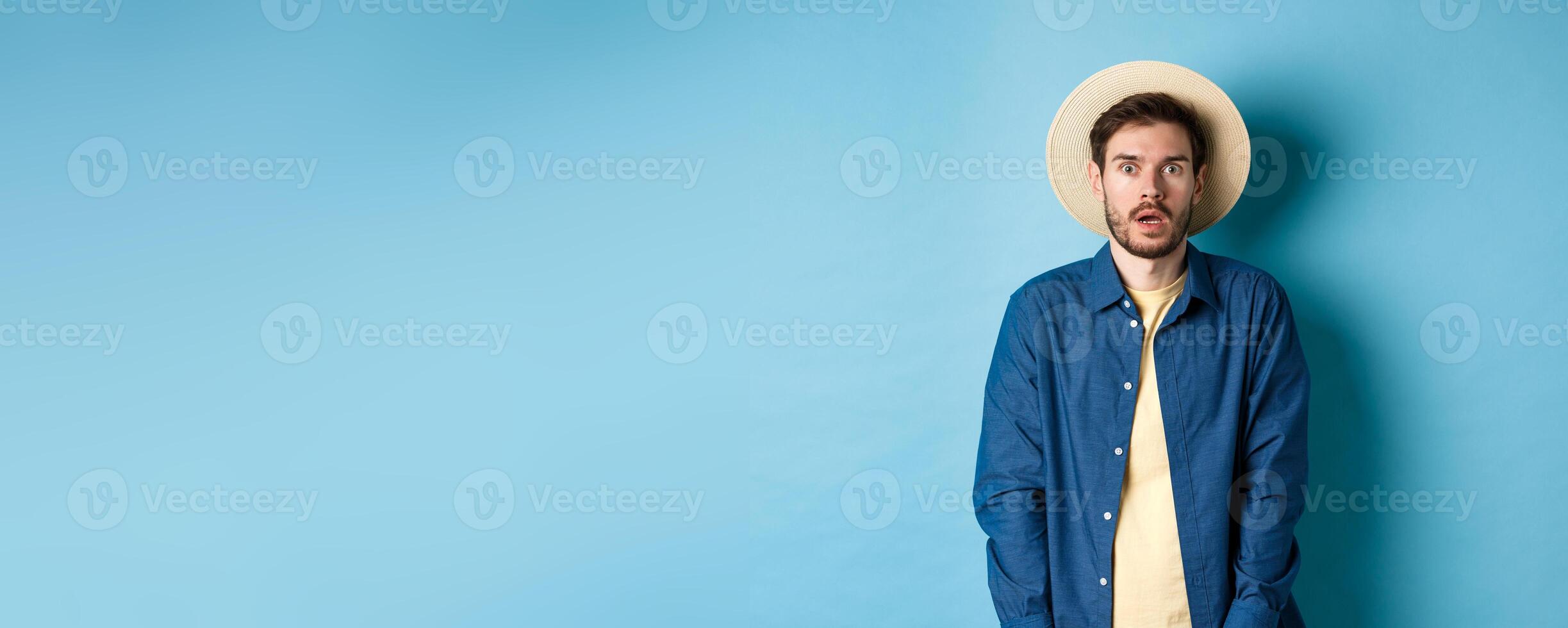 Young man tourist in summer hat drop jaw, staring at something shocking, standing on blue background photo