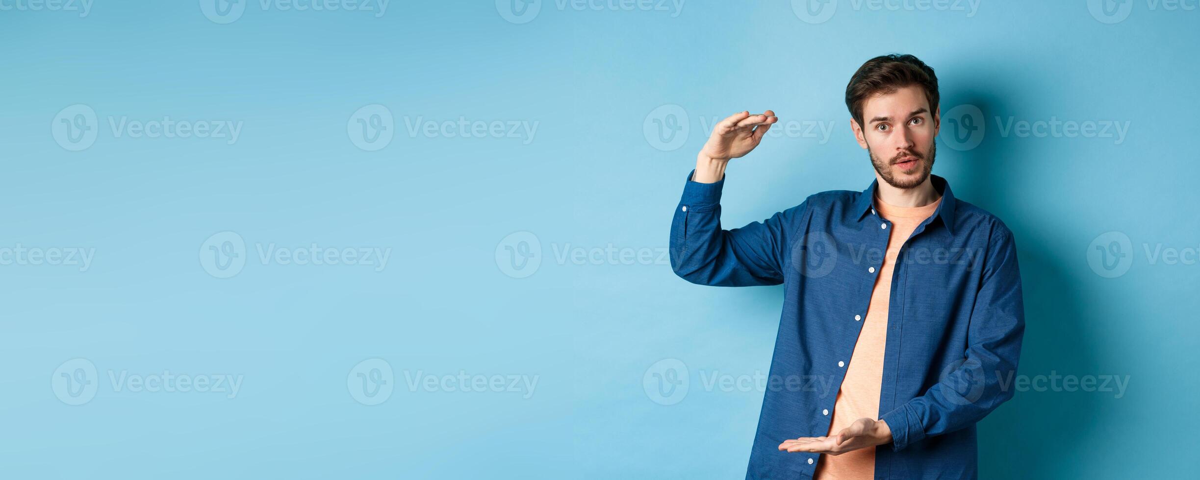 Modern guy showing big size, shaping large object and looking impressed at camera, standing on blue background photo