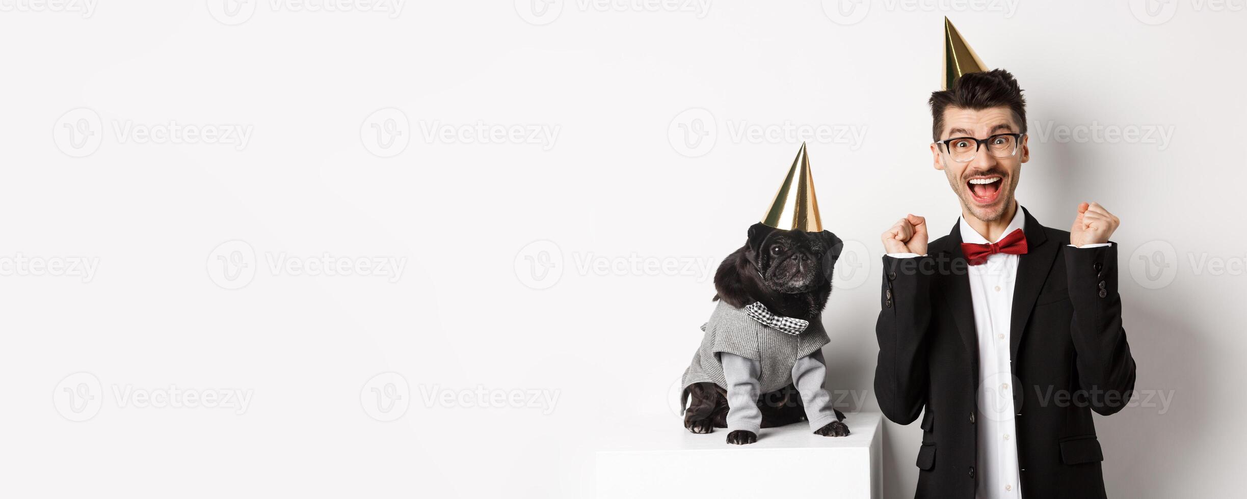 Cheerful young man shouting for joy, dog and owner wearing birthday party cones and celebrating, guy rejoicing and staring at camera, white background photo