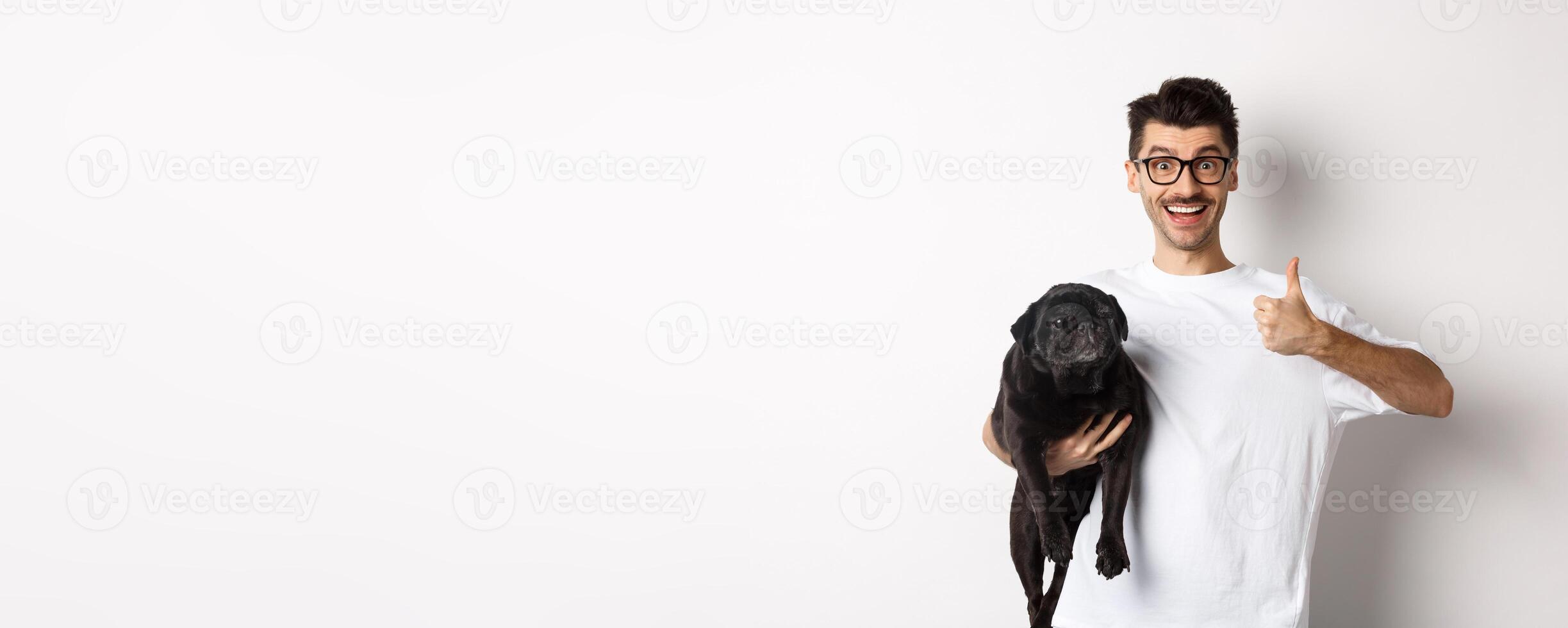 Cheerful handsome man holding dog and showing okay sign, approve or recommend product. Hipster guy carry cute black pug and looking satisfied, white background photo