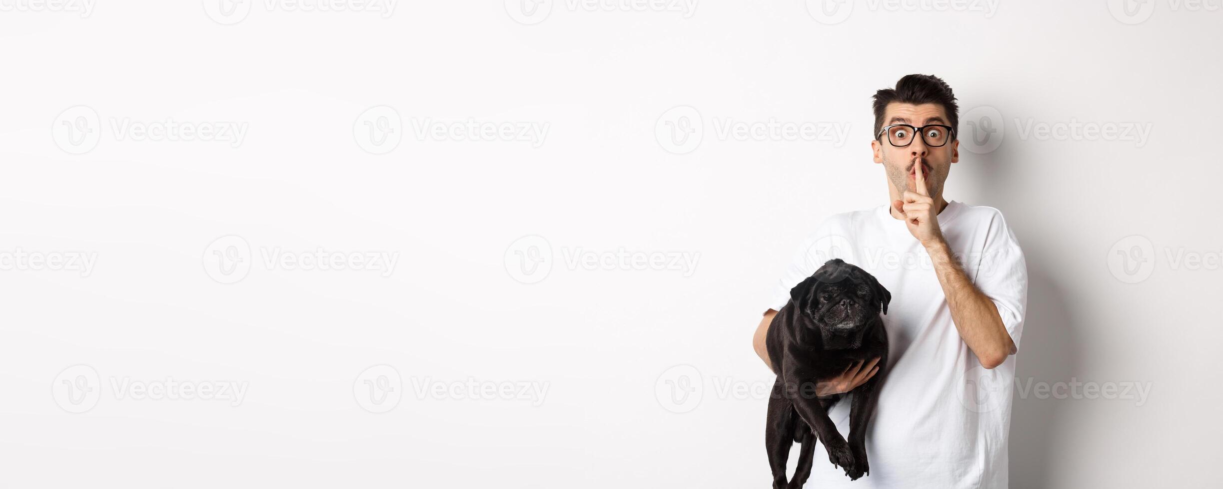 Image of funny young man holding cute black pug dog and shushing at camera, showing hush sign and staring at camera, keeping a secret, standing over white background photo