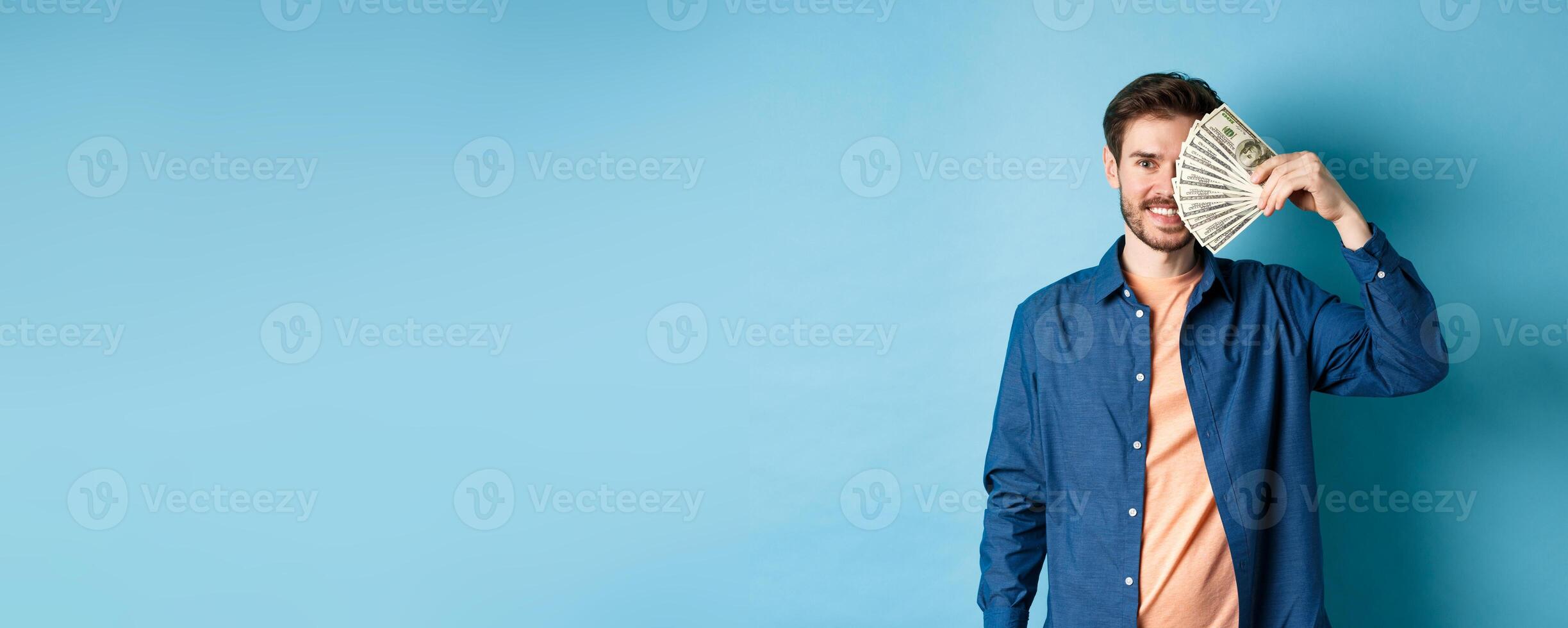 Happy young guy covering half of face with dollars and smiling, winning prize money, standing on blue background photo