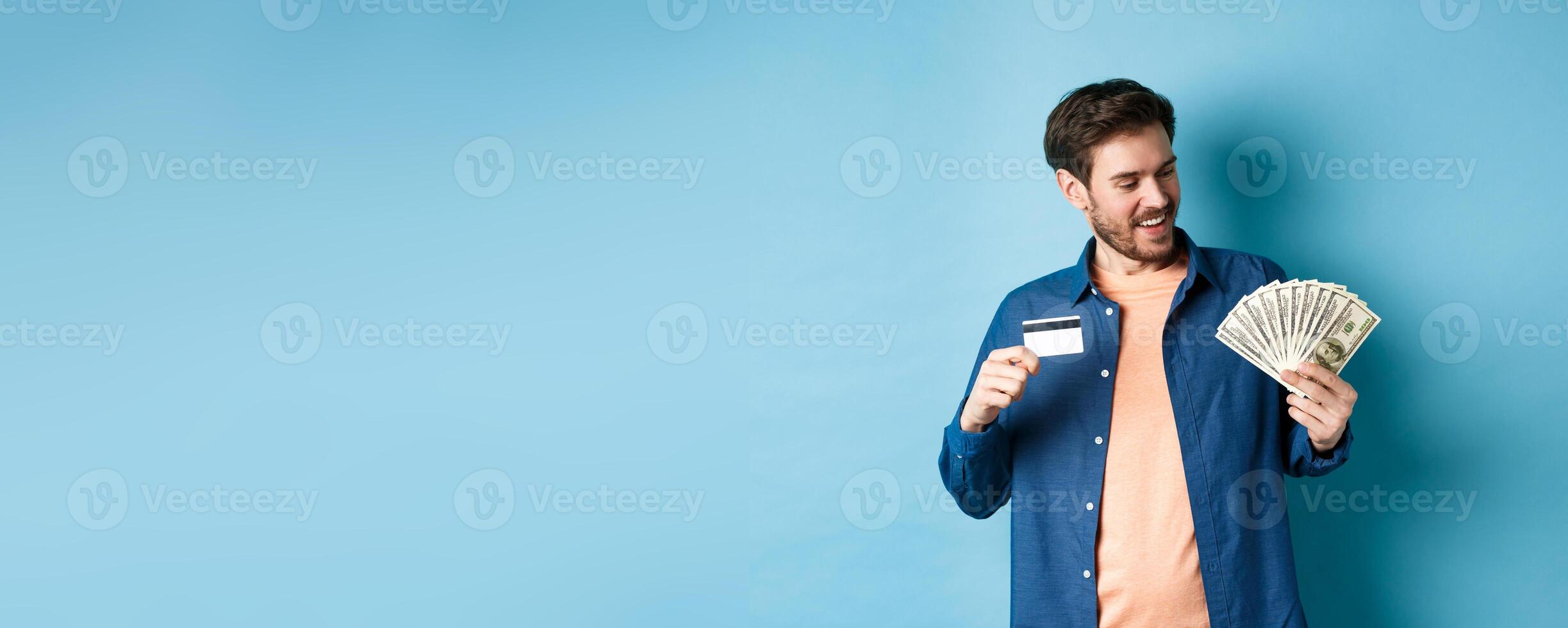 Smiling modern guy looking at cash and showing plastic credit card, standing on blue background photo