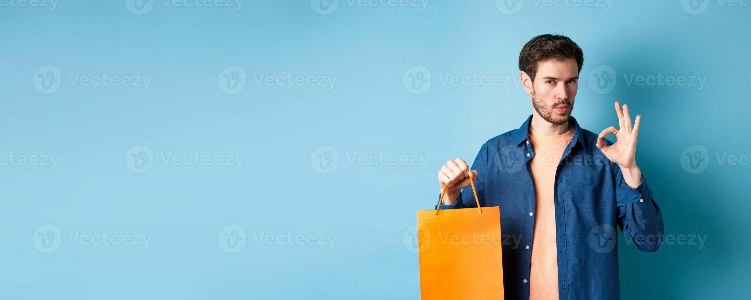 Handsome young man recommending store, showing orange shopping bag and OK sign, standing on blue background photo