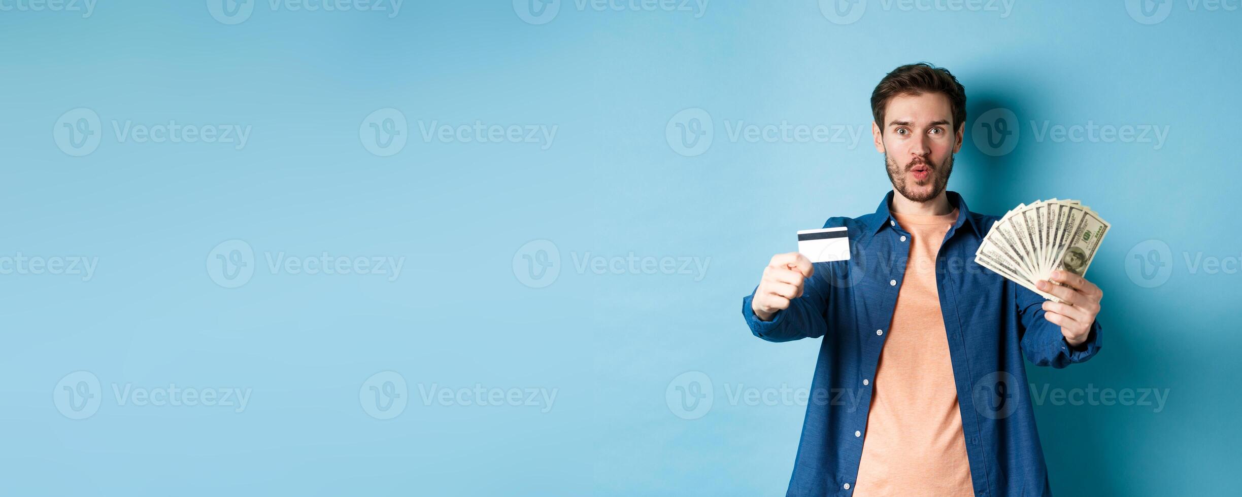 Excited modern guy showing money and plastic credit card, saying wow amazed, standing on blue background photo