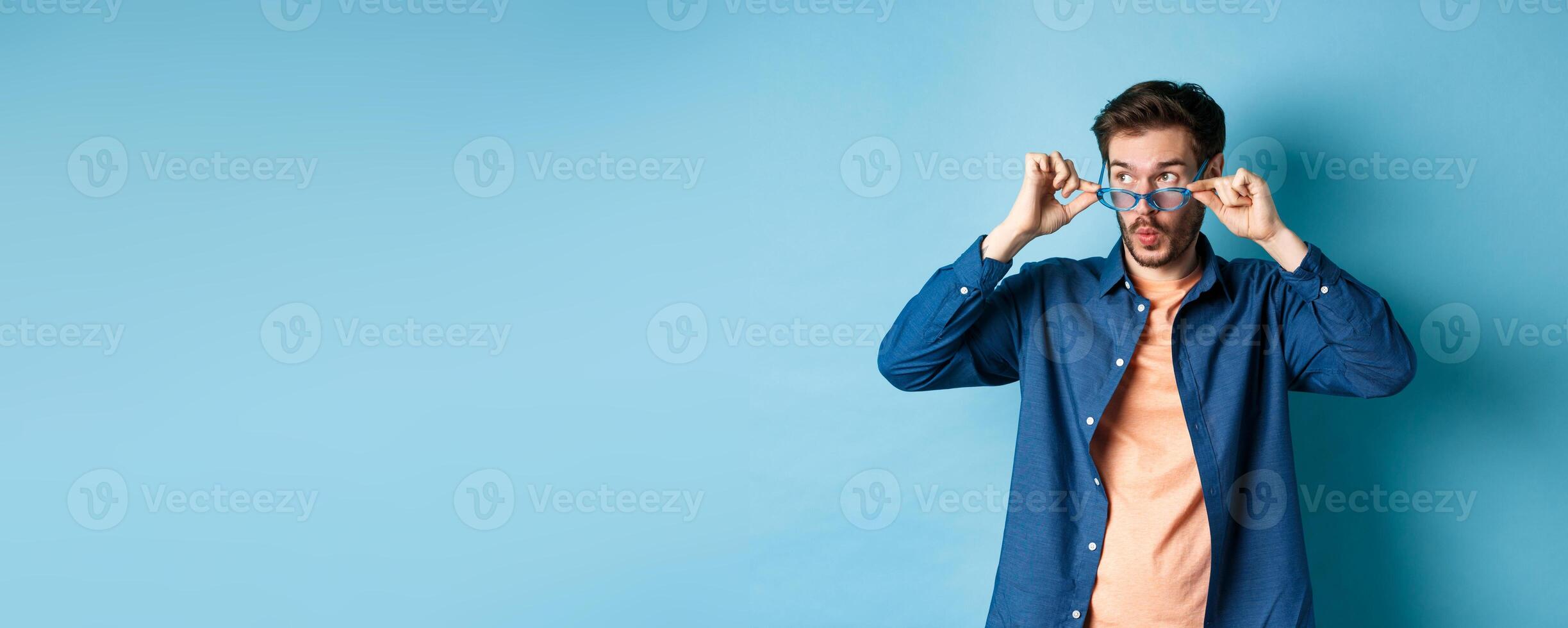 Image of caucasian guy takes-off sunglasses and looking aside with surprised face, checking out empty space for logo, standing on blue background photo