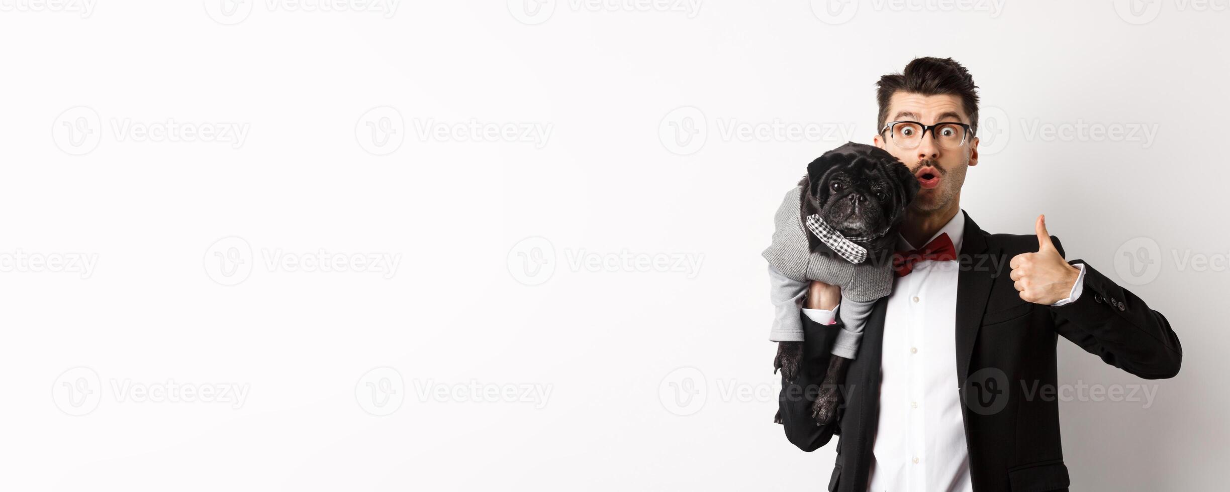 Happy young hipster in suit and glasses, showing thumb-up, holding cute black dog on shoulder, love his pug, standing over white background photo