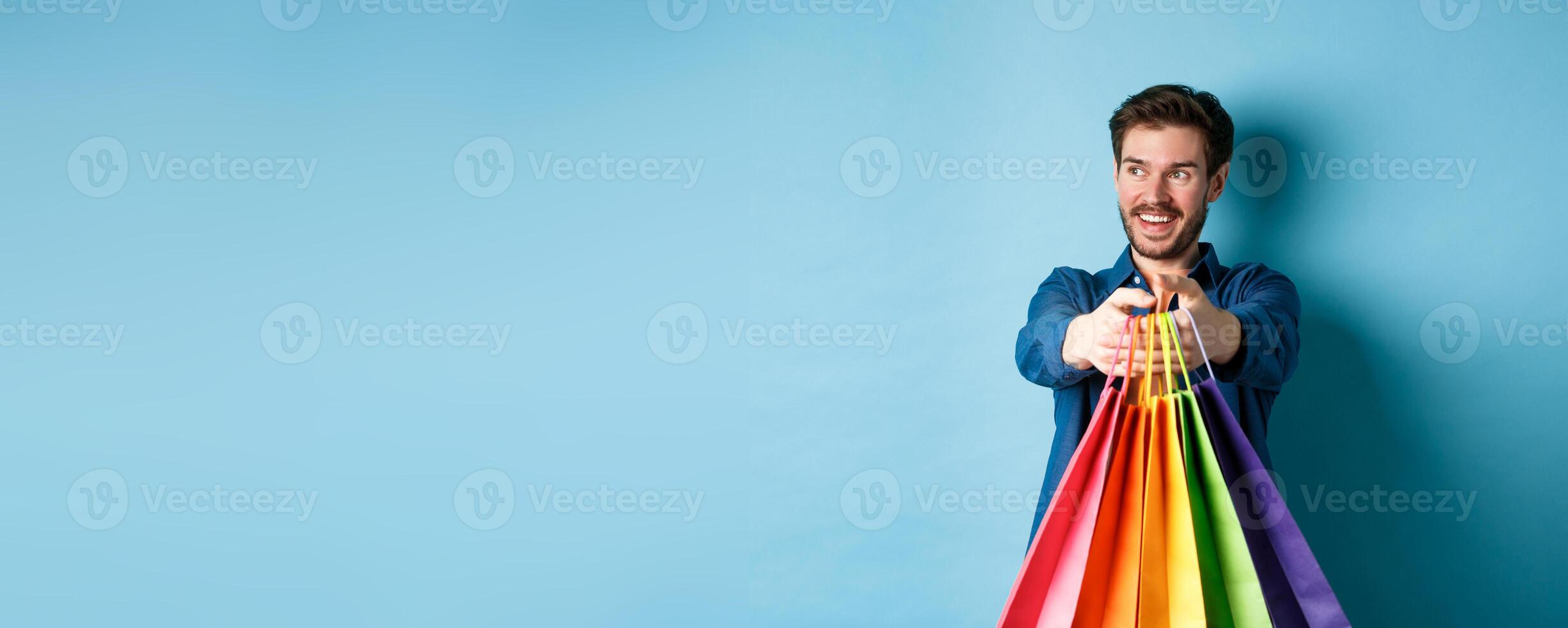 Handsome happy guy smiling, stretch out hands with shopping bags and looking aside at empty space, standing on blue background photo