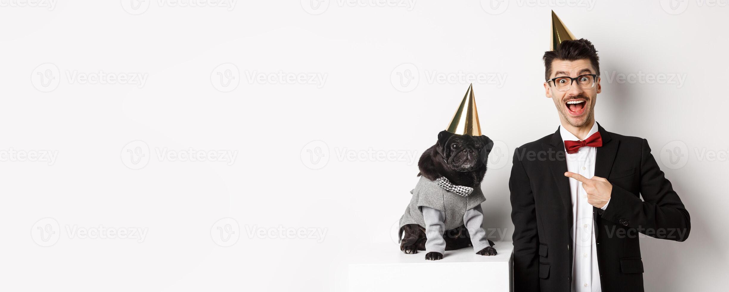 Happy dog owner and black pug wearing birthday party cones, man pointing at pug, standing over white background photo