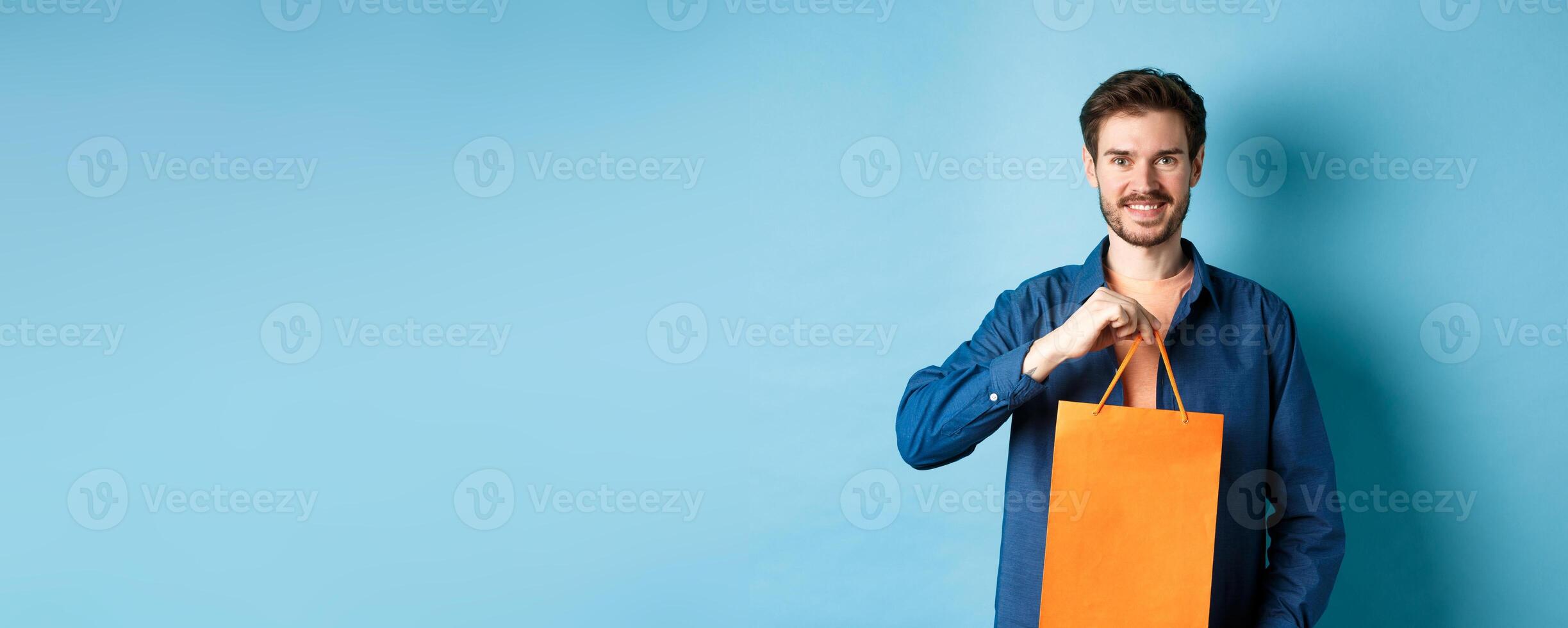 Handsome young man with beard, smiling and showing shopping bag, buying something in store, standing on blue background photo