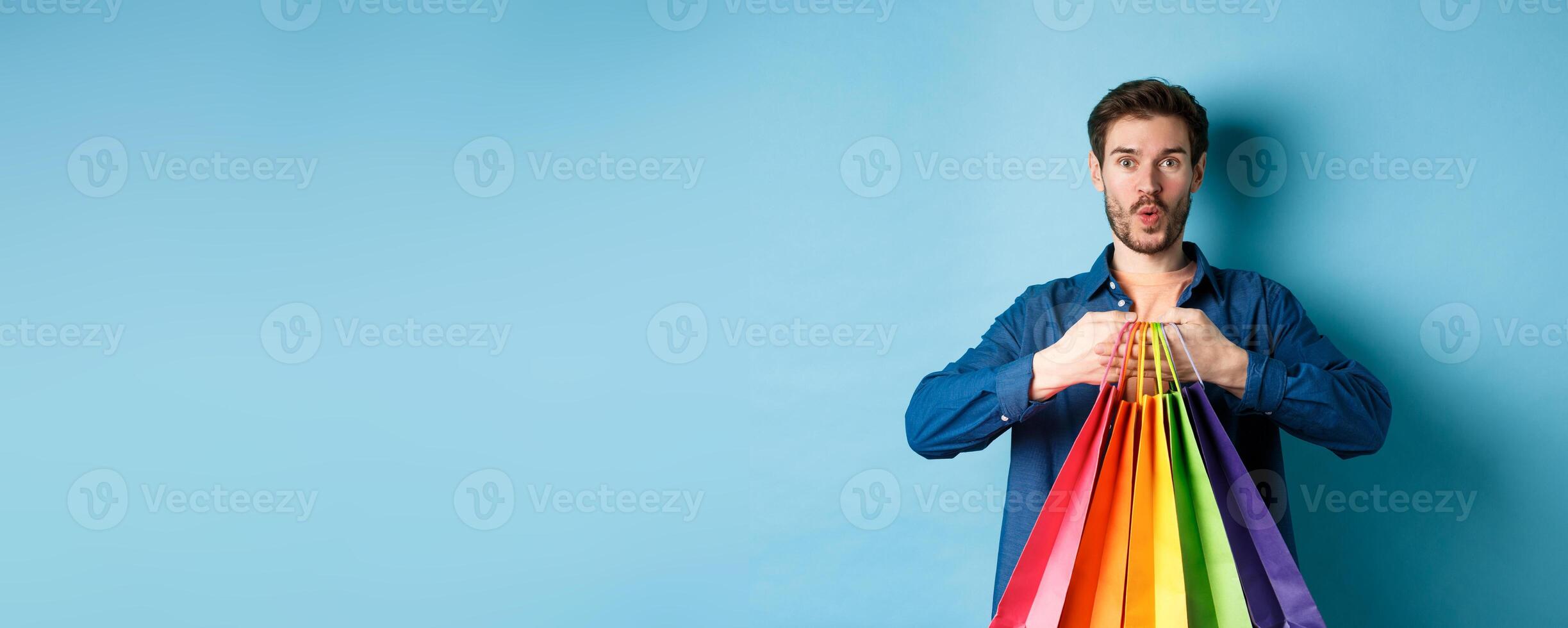 Excited shopper holding packages in paper bags and smiling amused, standing on blue background photo