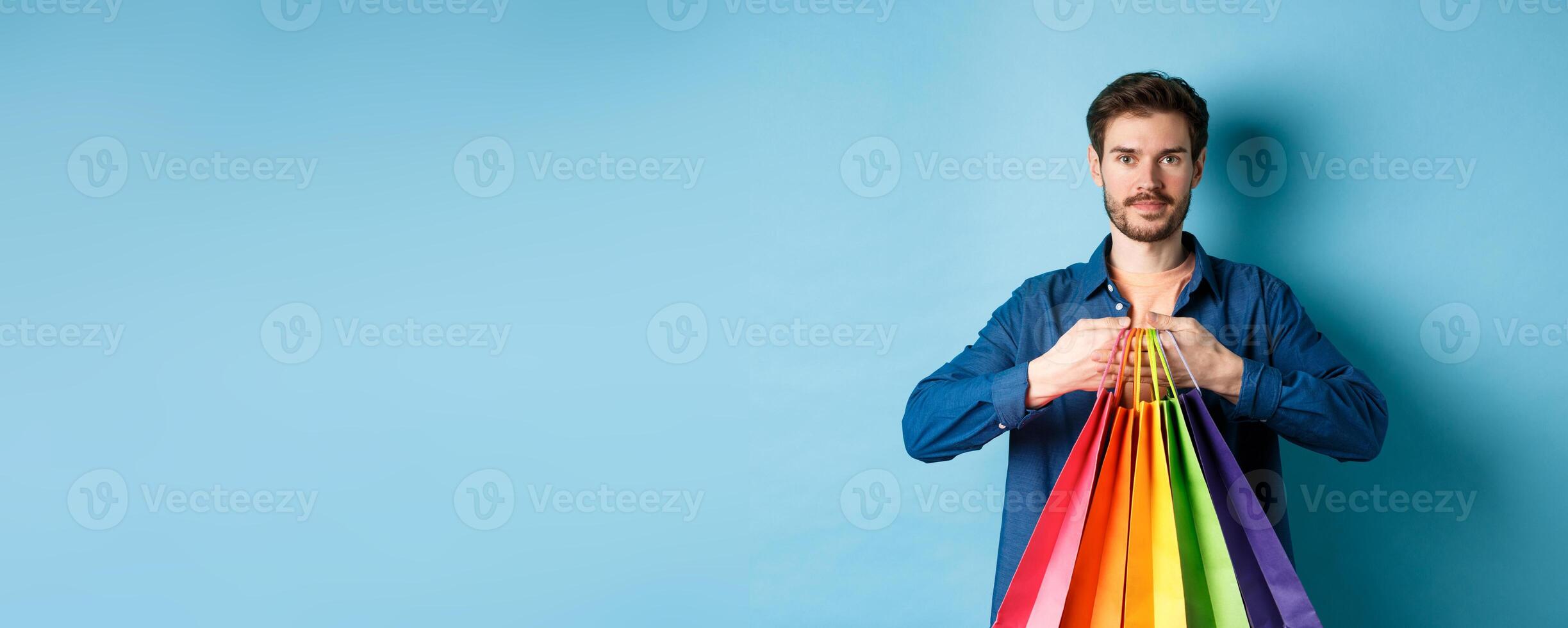 Modern guy with beard, holding packages from shop and smiling, standing on blue background photo