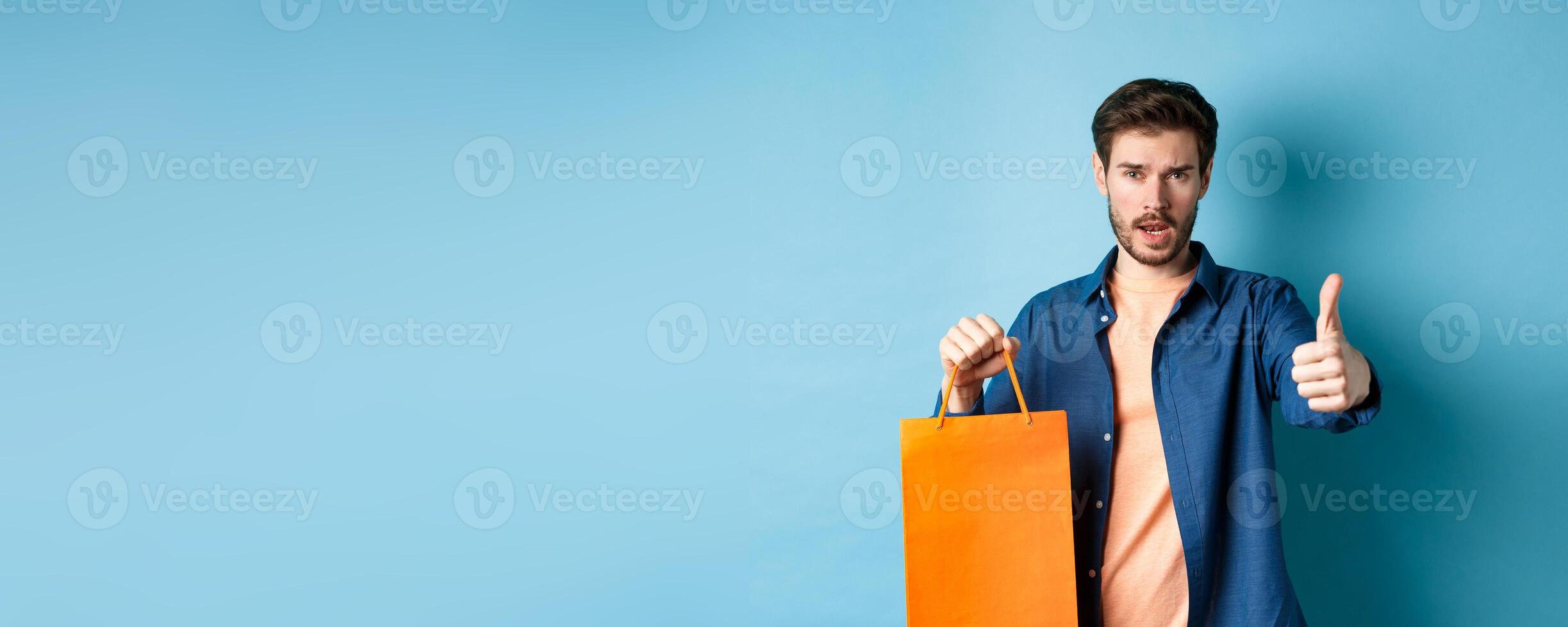 Cool guy showing thumb up and holding orange shopping bag, standing on blue background photo