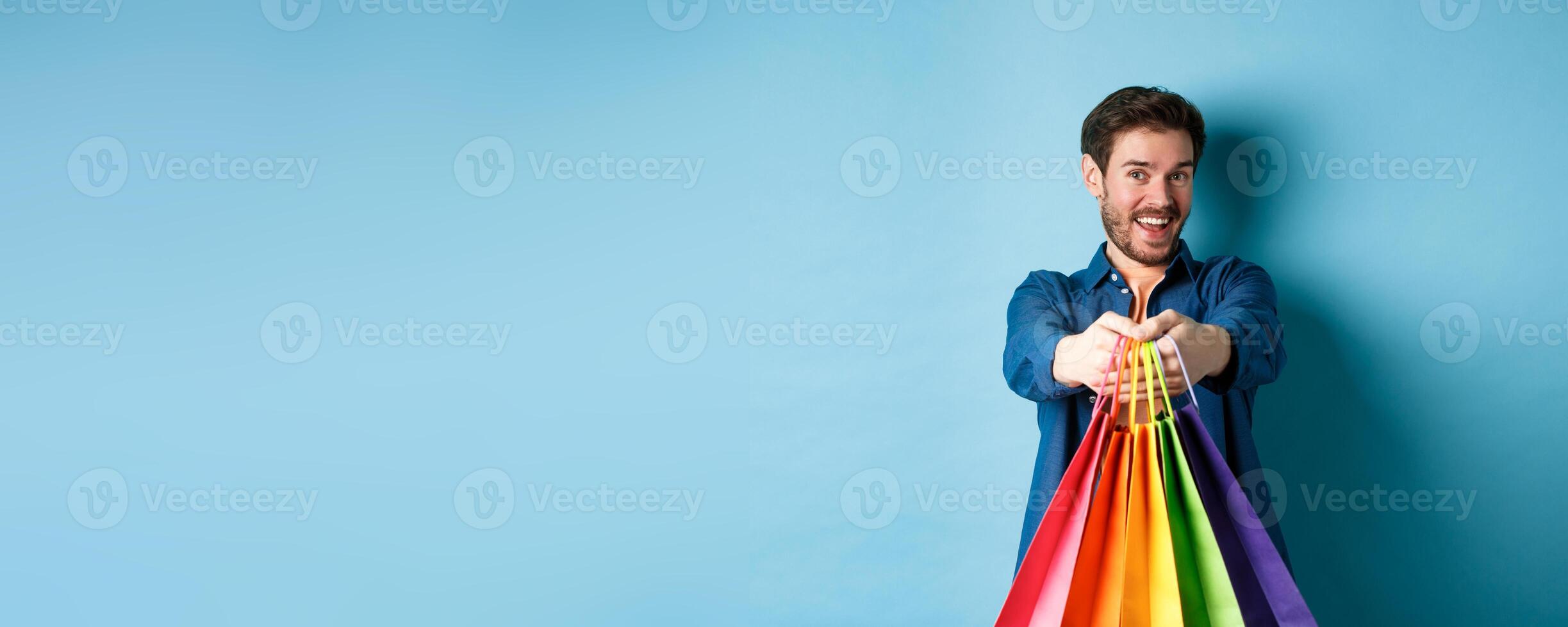 Cheerful young guy stretch out hands with shopping bags and smiling, giving you gifts, standing on blue background photo