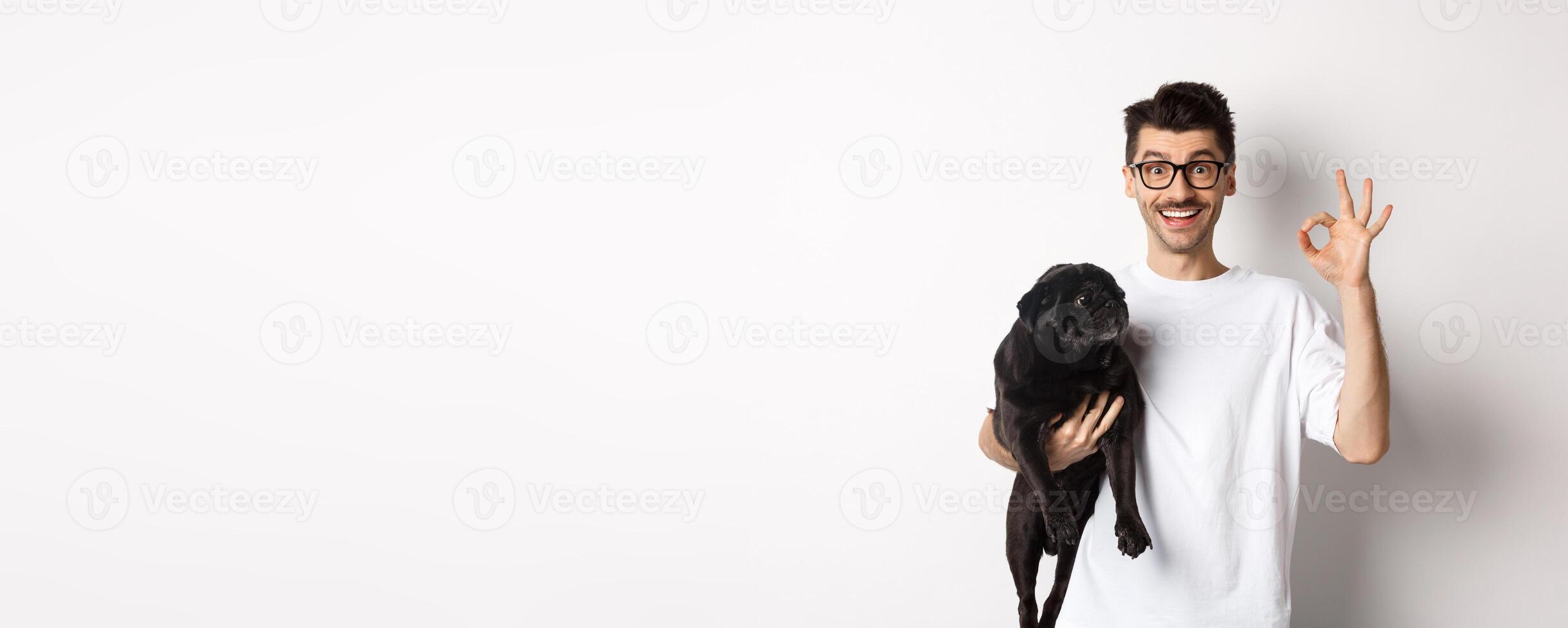 Cheerful handsome man holding dog and showing okay sign, approve or recommend pet related product, standing over white background photo