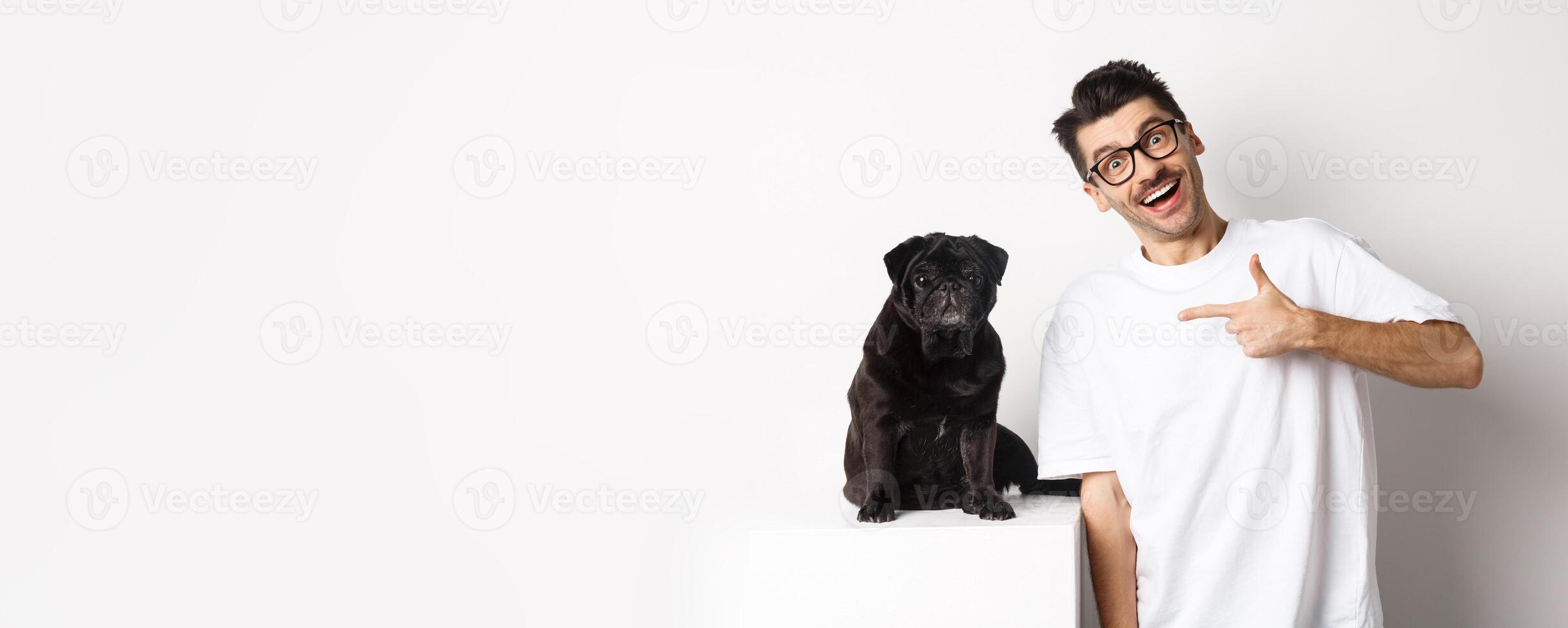 Happy young man showing his cute dog, pointing finger at black pug and smiling, standing over white background photo